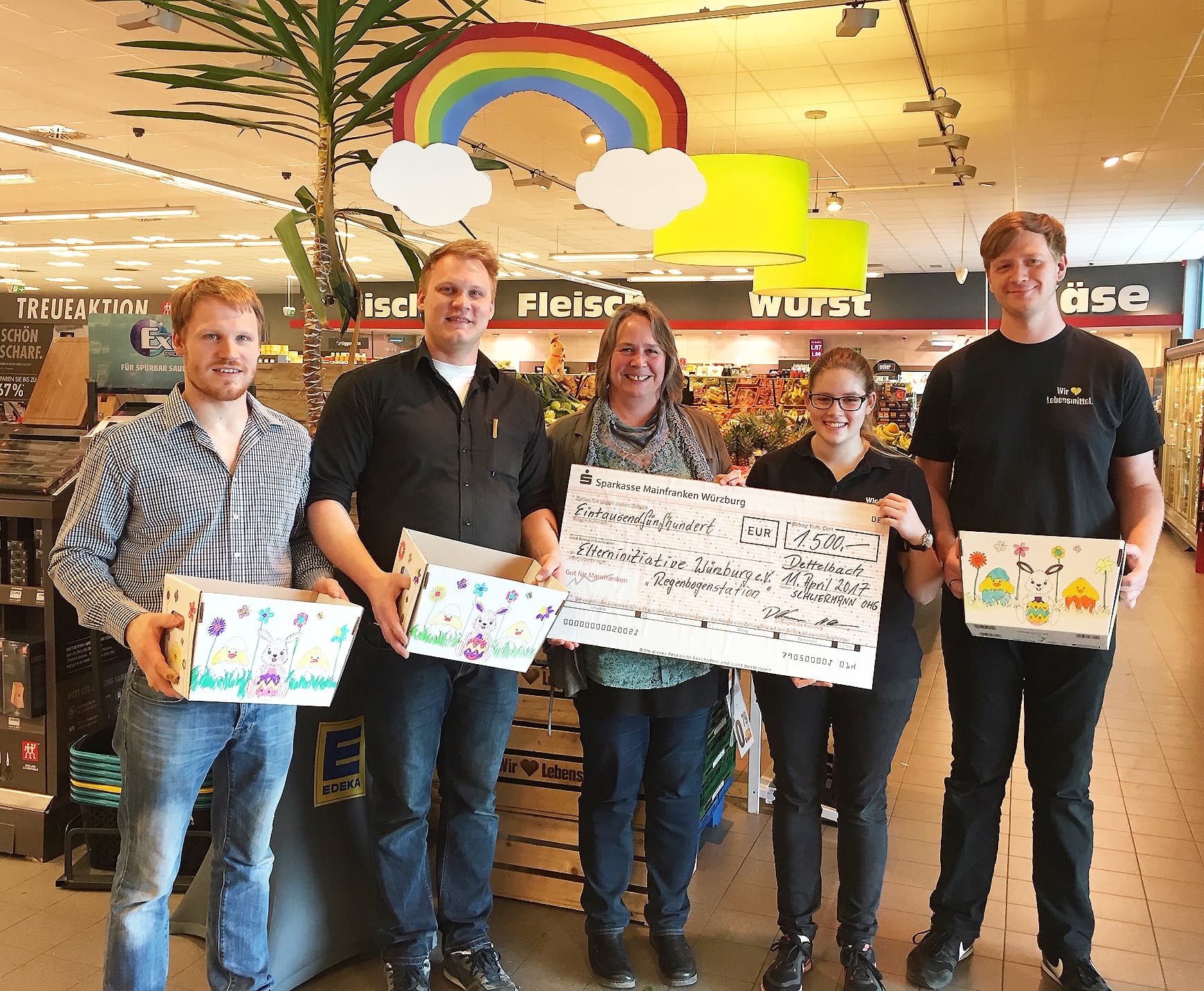 Fünf Personen stehen lächelnd in einem EDEKA-Markt. Sie halten einen Scheck und verzierte Pappkisten in den Händen.