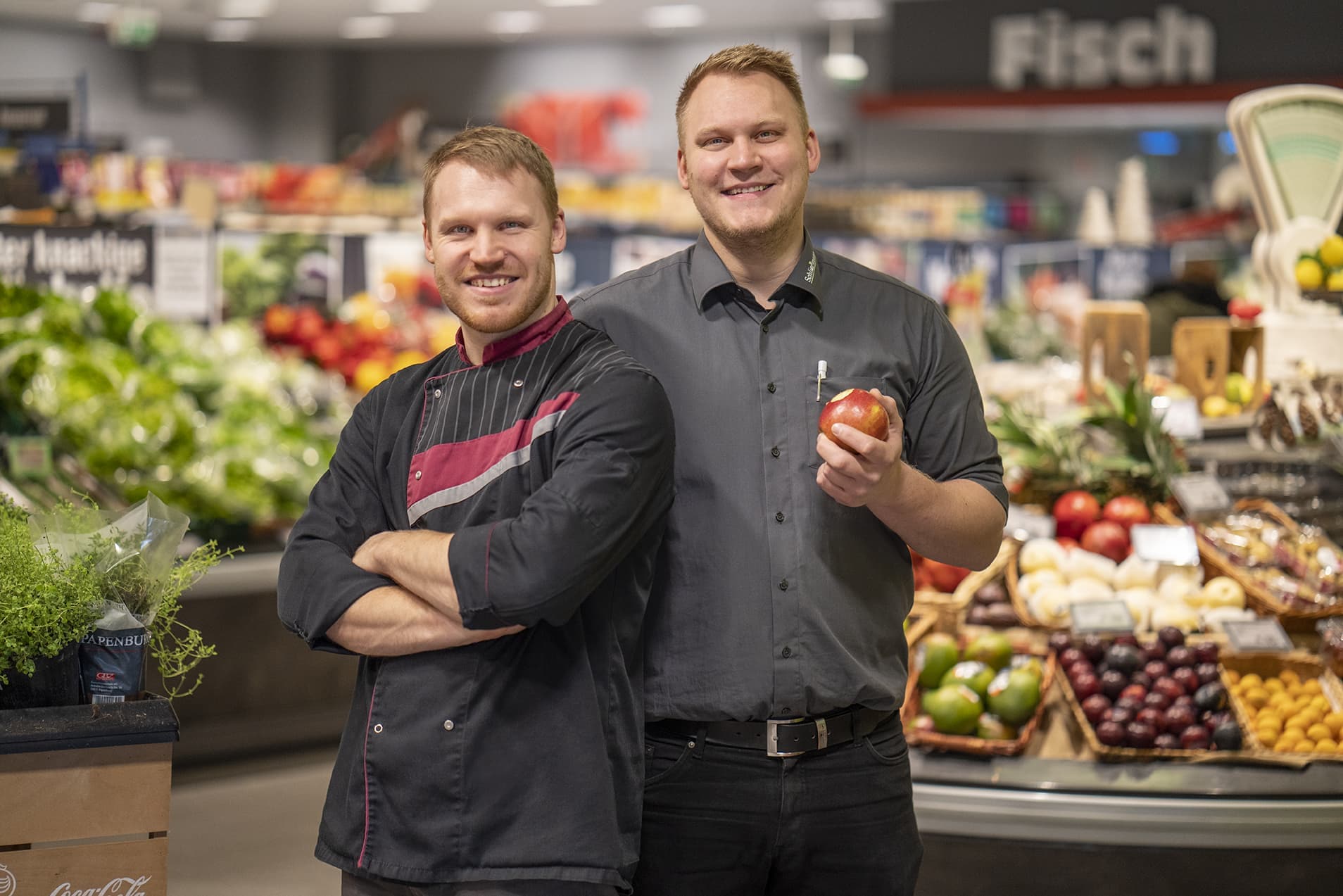 Die beiden jungen Marktleiter, einer mit verschränkten Armen, einer mit angebissenem Apfel in der Hand, lächelnd in der Obst- und Gemüseabteilung eines EDEKA-Markts.