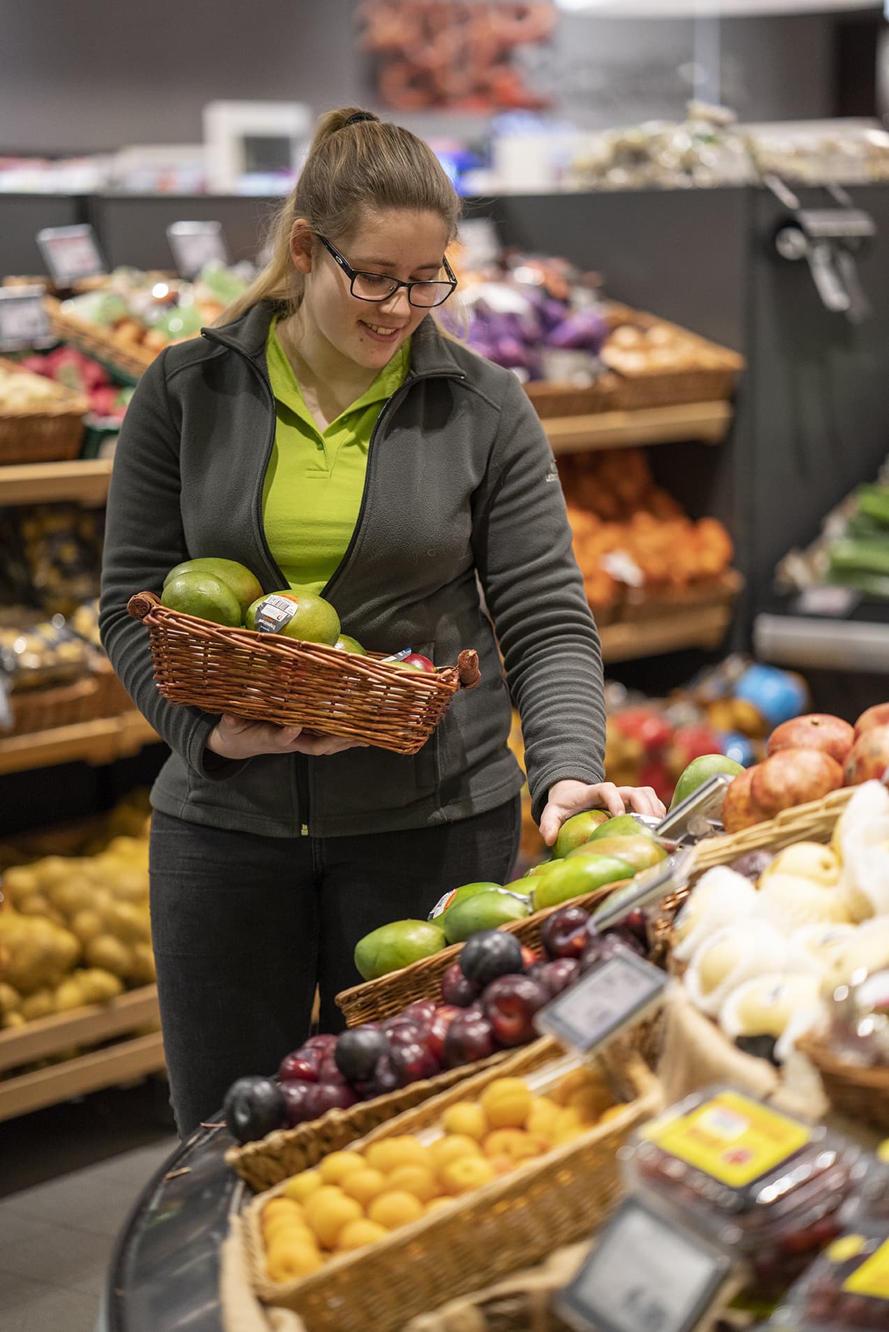 Eine Mitarbeiterin in der Obst- und Gemüseabteilung legt Obst in ein Fach. Sie lächelt.