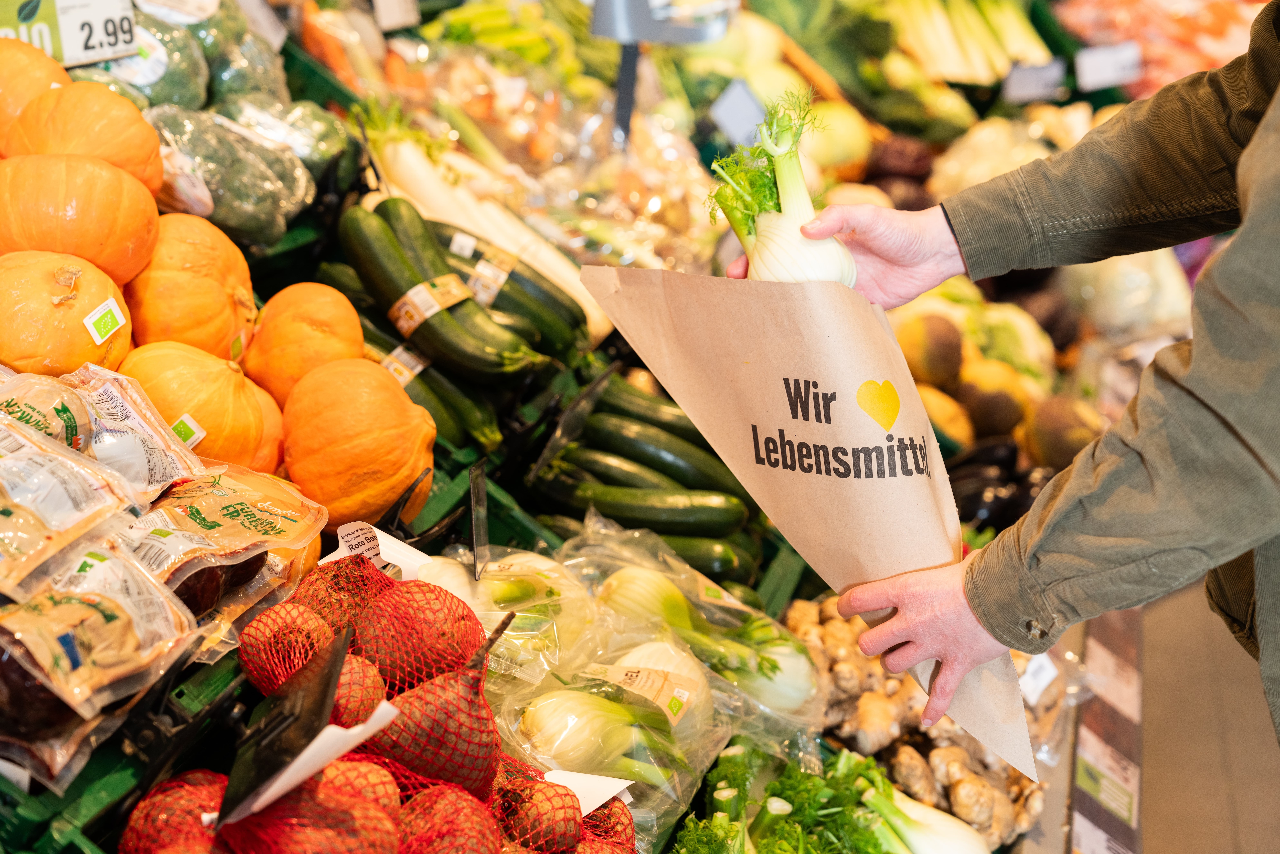 Eine Hand legt einen Fenchel in eine Papiertüte mit der Aufschrift: "Wir lieben Lebensmittel"