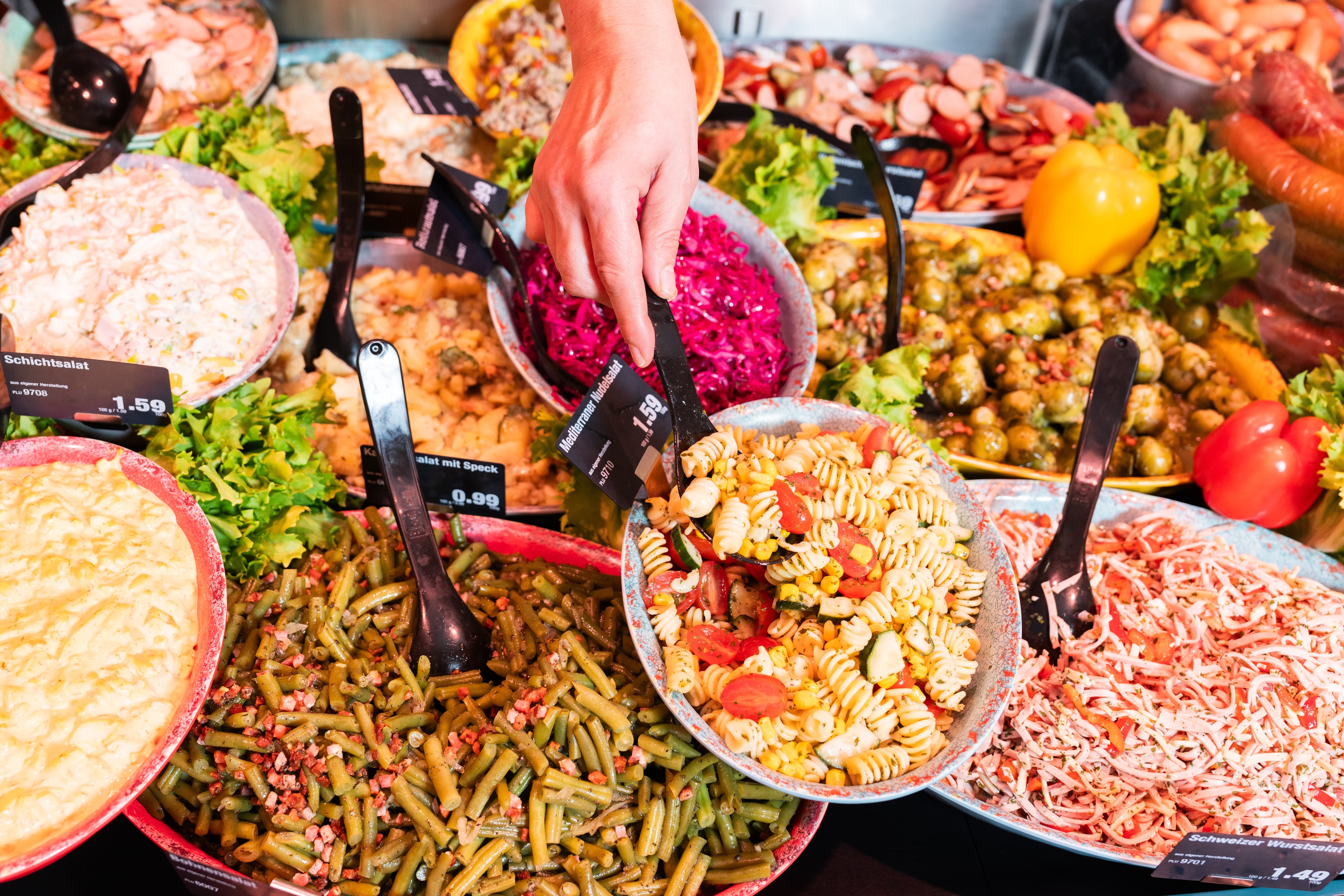 Eine Hand steckt einen Löffeln in eine Schüssel mit Nudelsalat. Diese Schüssel steht zusammen mit anderen Schüsseln voller bunter Salate in der Auslage einer Frischetheke.