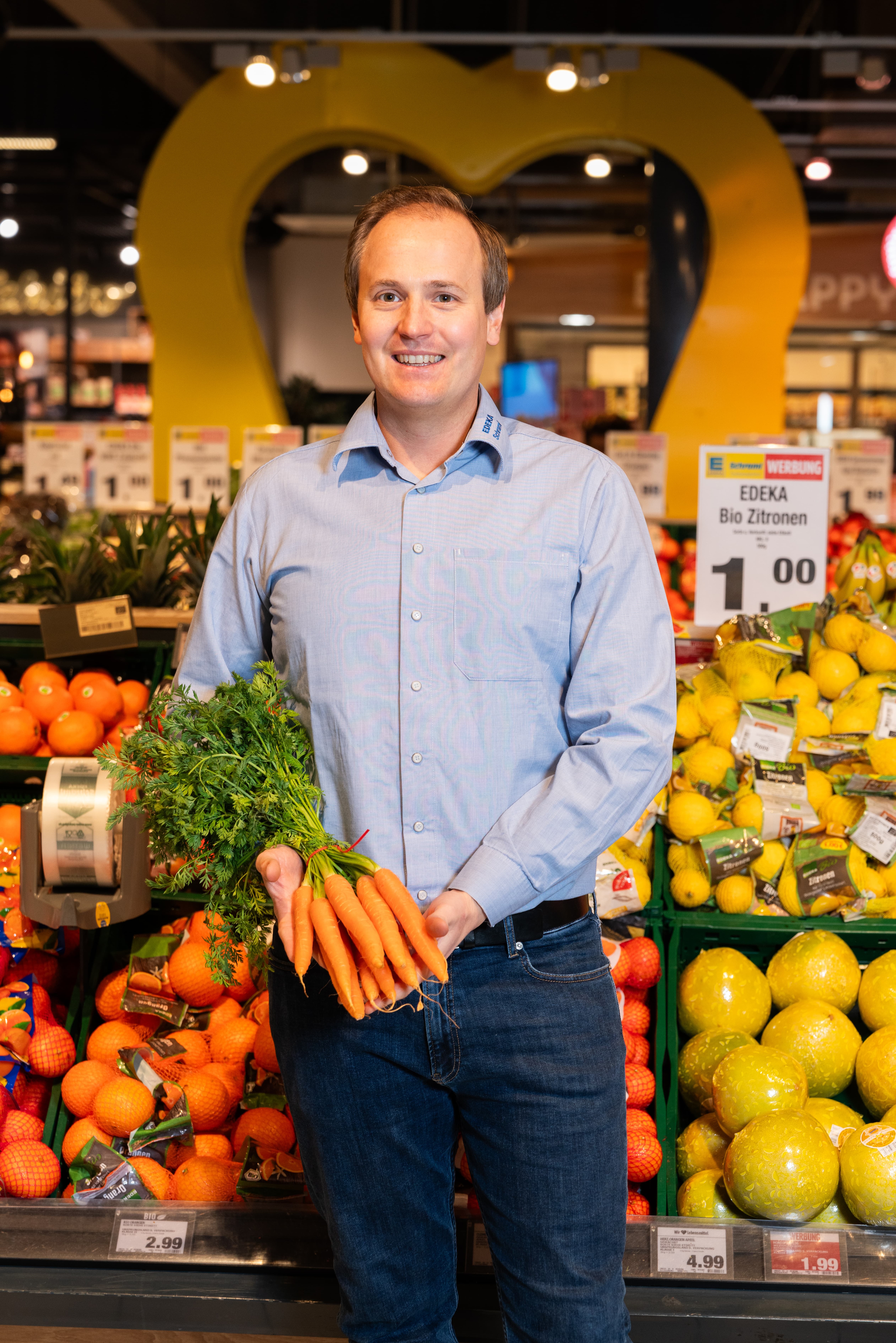 Der lächelnde Marktinhaber steht in der Obst- und Gemüseabteilung und hält ein Bund Möhren in den Händen.