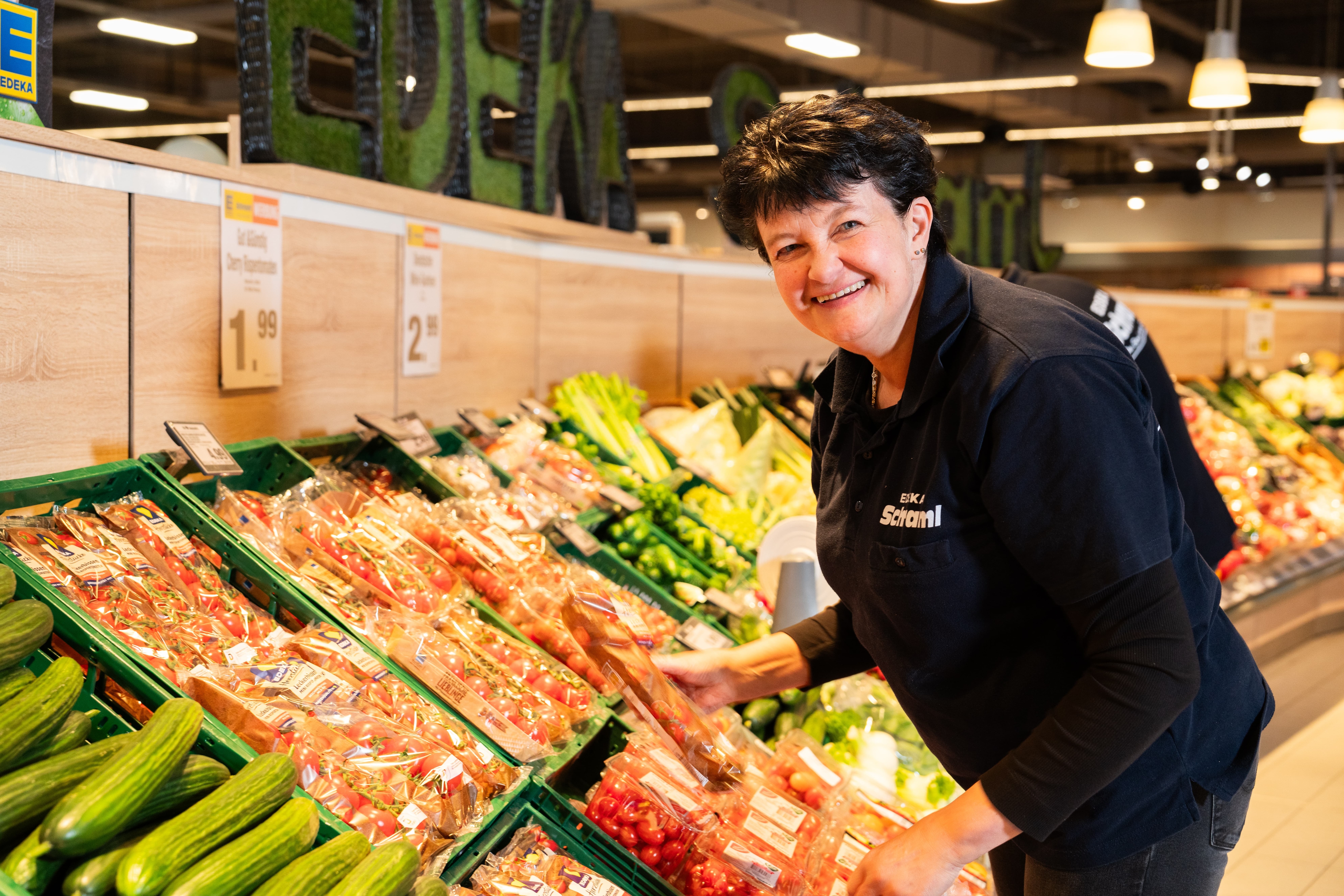 Eine lächelnde EDEKA-Mitarbeiterin sortiert Tomaten in die Auslage einer Gemüseabteilung.