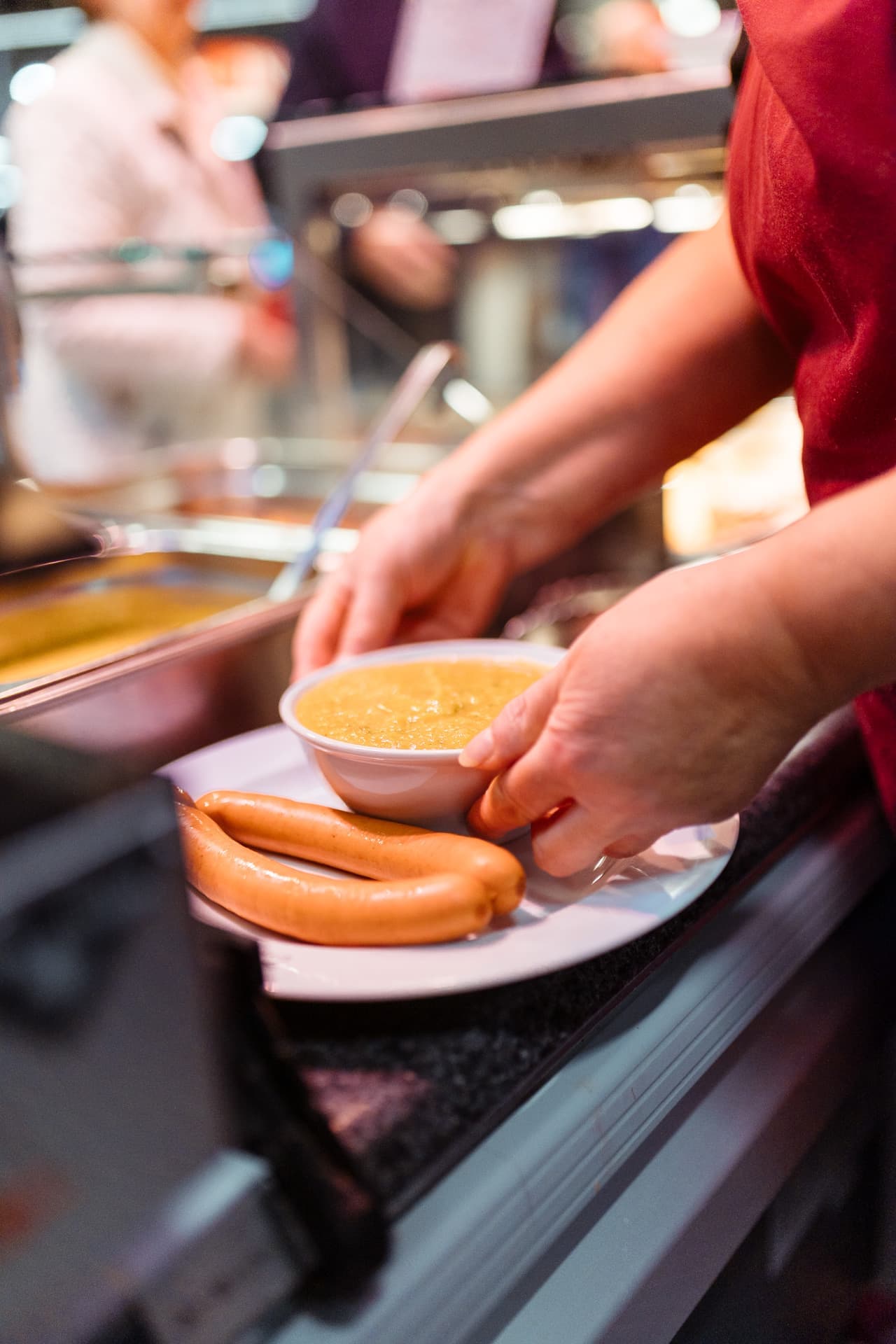 Nahaufnahme von Händen, die eine Schüssel Suppe auf einen Teller stellen, auf dem bereits zwei Würstchen liegen.