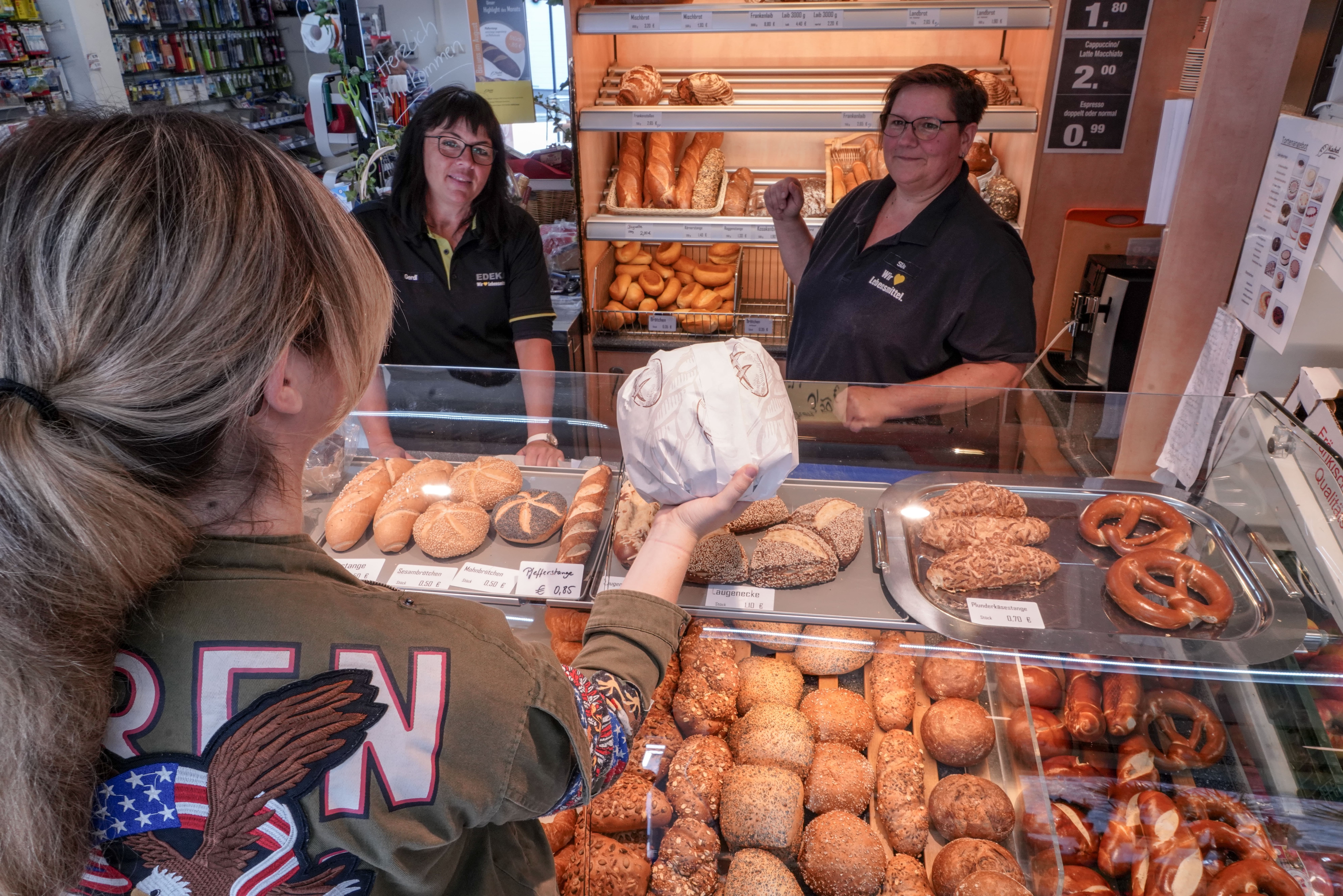 Blick von oben auf die Bäckereitheke eines EDEKA-Markts. Zwei Mitarbeiterinnen stehen lächelnd dahinter, eine Kundin nimmt ein verpacktes Brot entgegen.