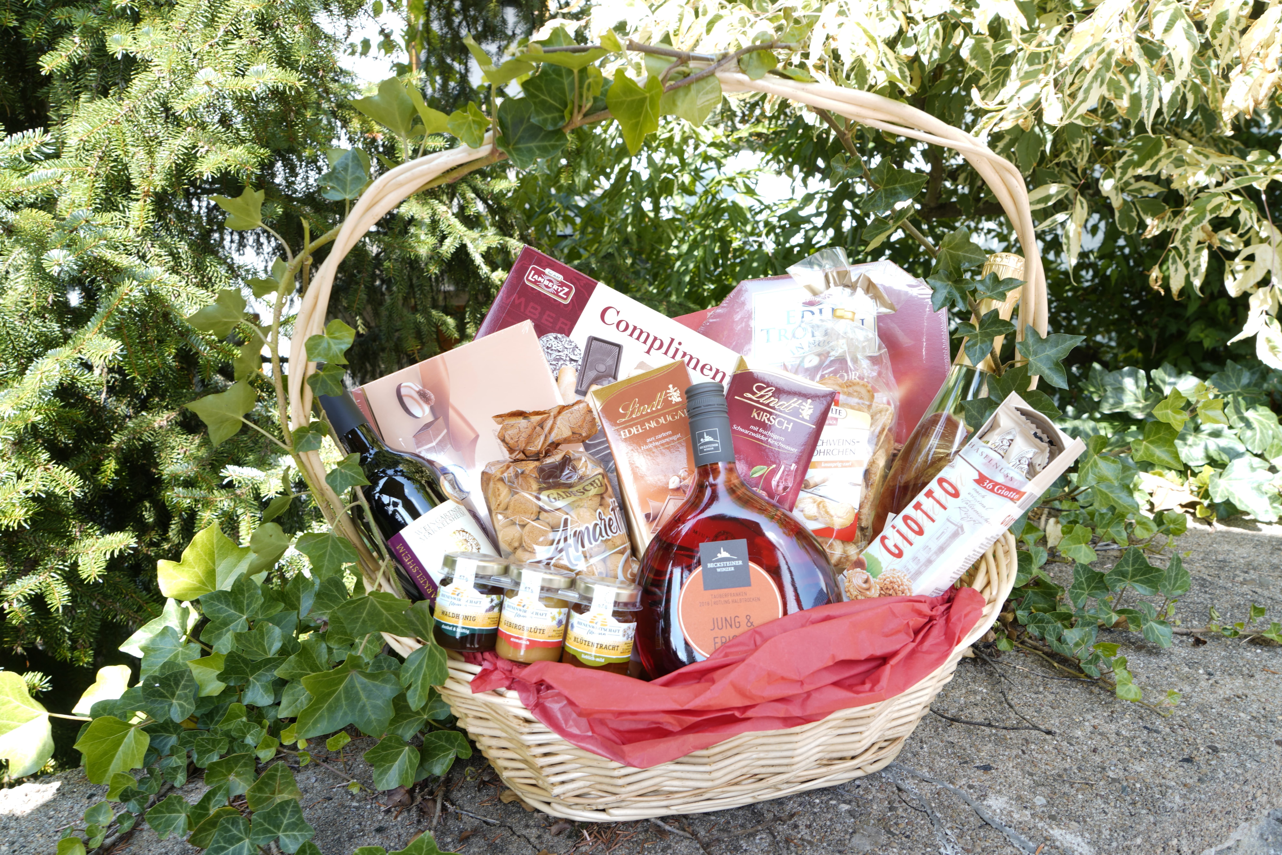 Ein Präsentkorb mit verschiedenen Genussmitteln steht vor einem grünen Gebüsch.
