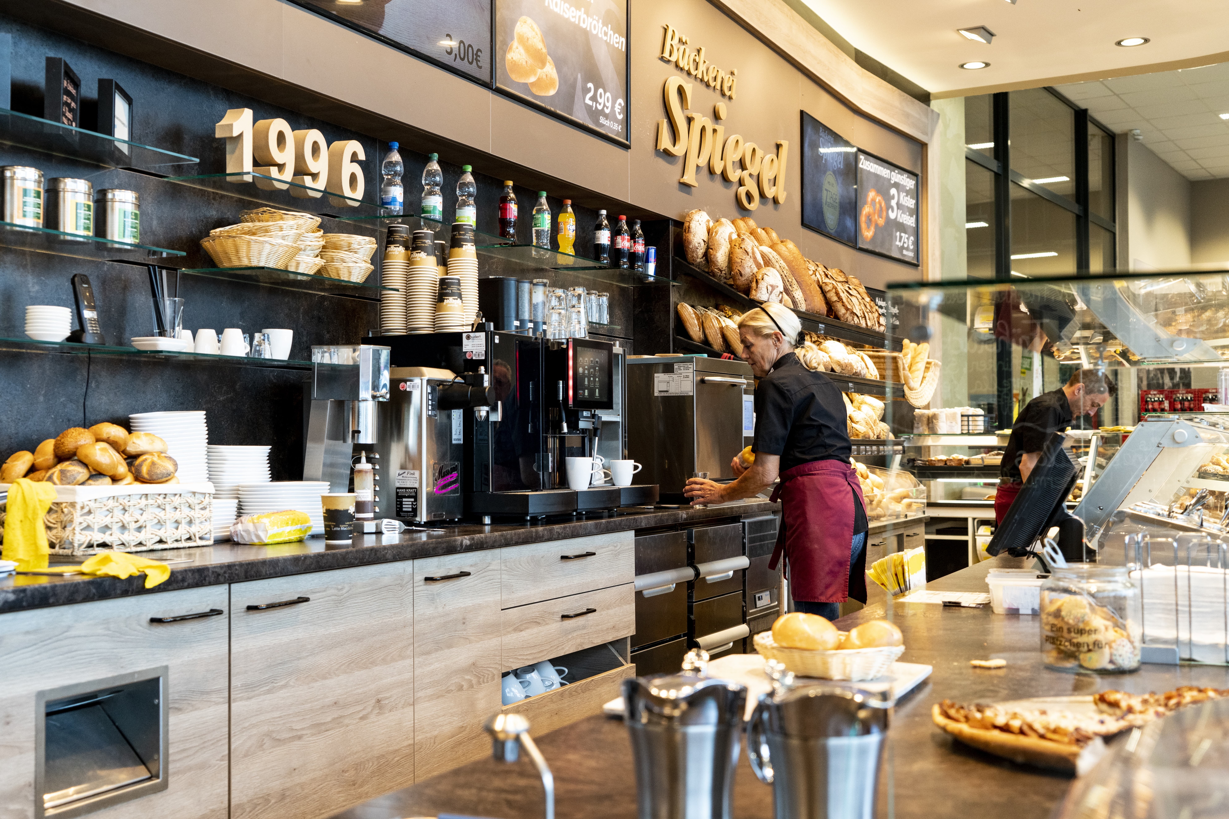 Die Bäckerei in diesem EDEKA-Markt, betitelt mit „Bäckerei Spiegel“. Man sieht den Arbeitsbereich hinter der Theke, wo zwei Mitarbeitende arbeiten.