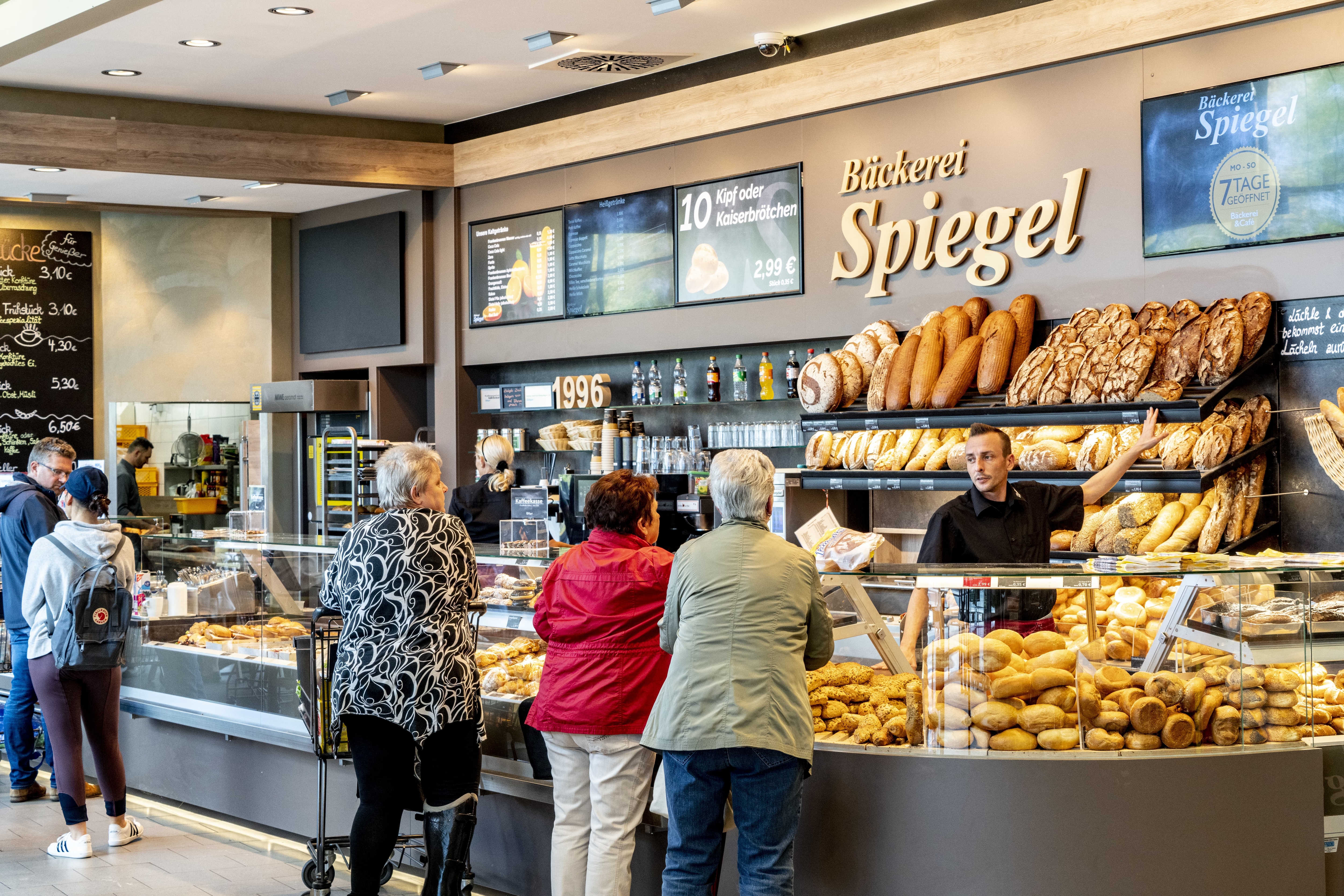 Die Bäckerei in diesem EDEKA-Markt, betitelt mit „Bäckerei Spiegel“ und verschiedenen Backwaren, belegt und unbelegt, in der Auslage. Kunden stehen vor der Auslage und lassen sich von einem Mitarbeiter beraten.