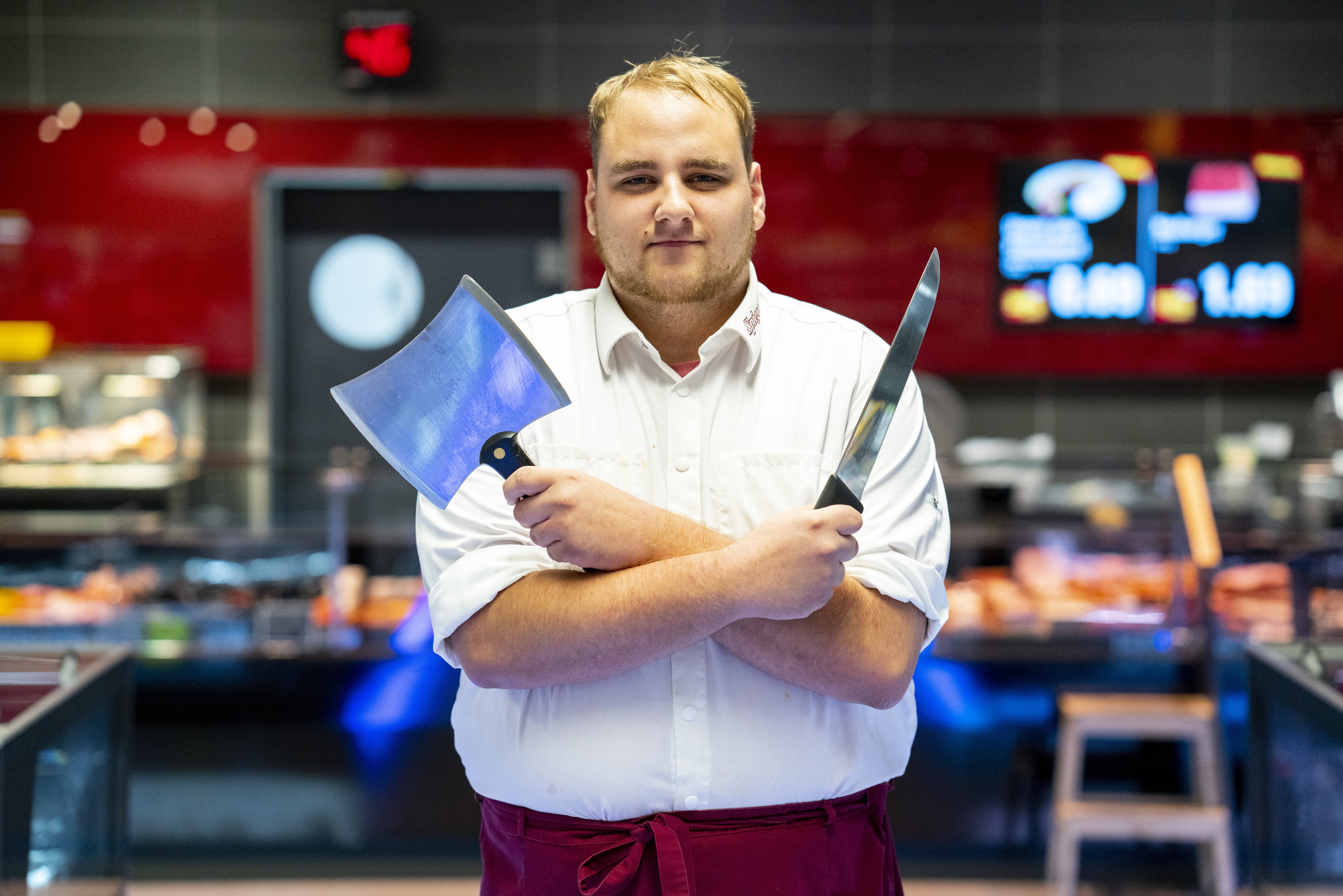 Ein junger Mitarbeiter steht vor der Bedientheke eines EDEKA-Markts. Er hält ein Metzgerbeil und ein Messer über Kreuz vor der Brust wie ein altertümlicher Krieger.