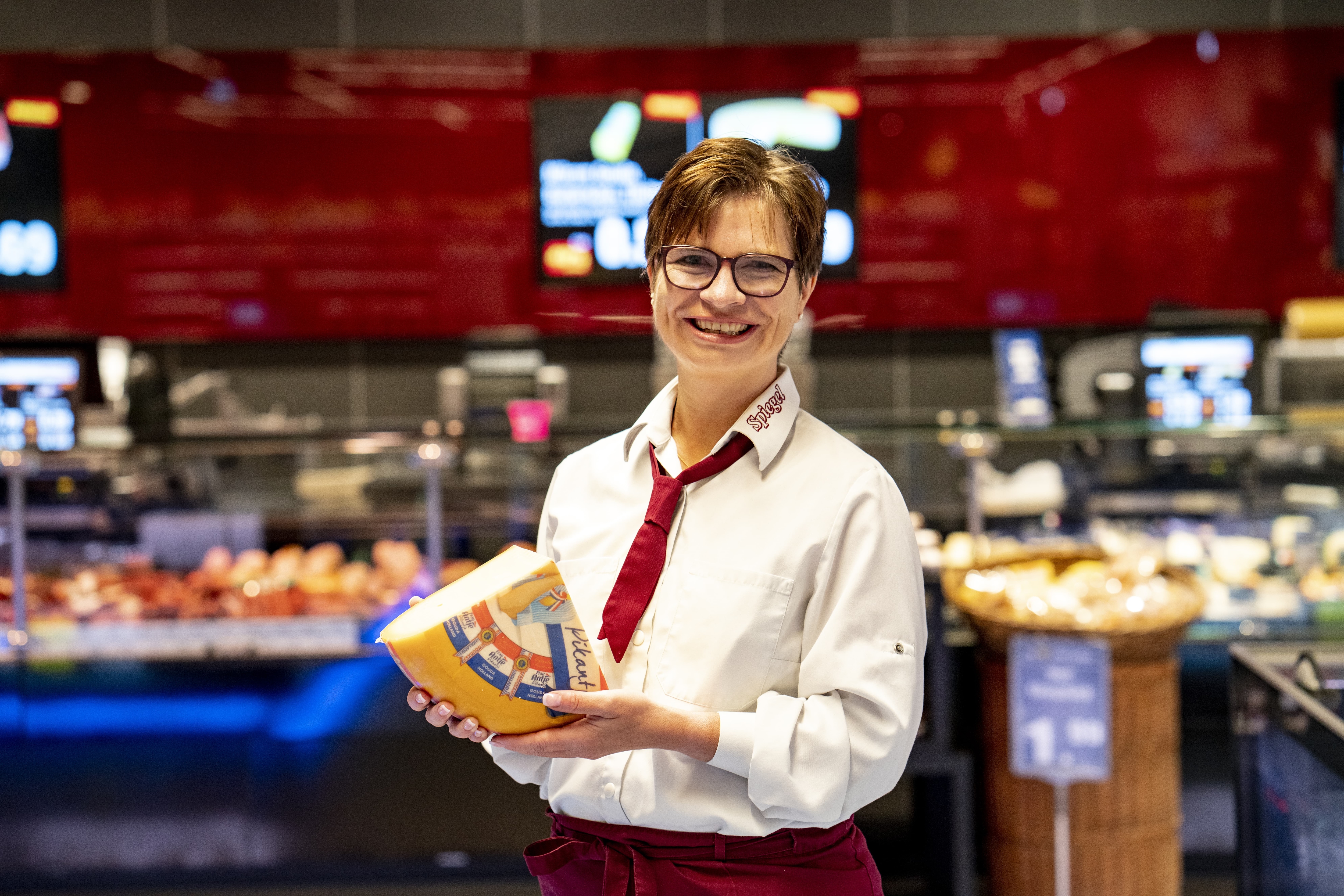 Eine Mitarbeiterin mit einem Viertellaib Käse in den Händen steht lächelnd vor der Bedientheke eines EDEKA-Markts.