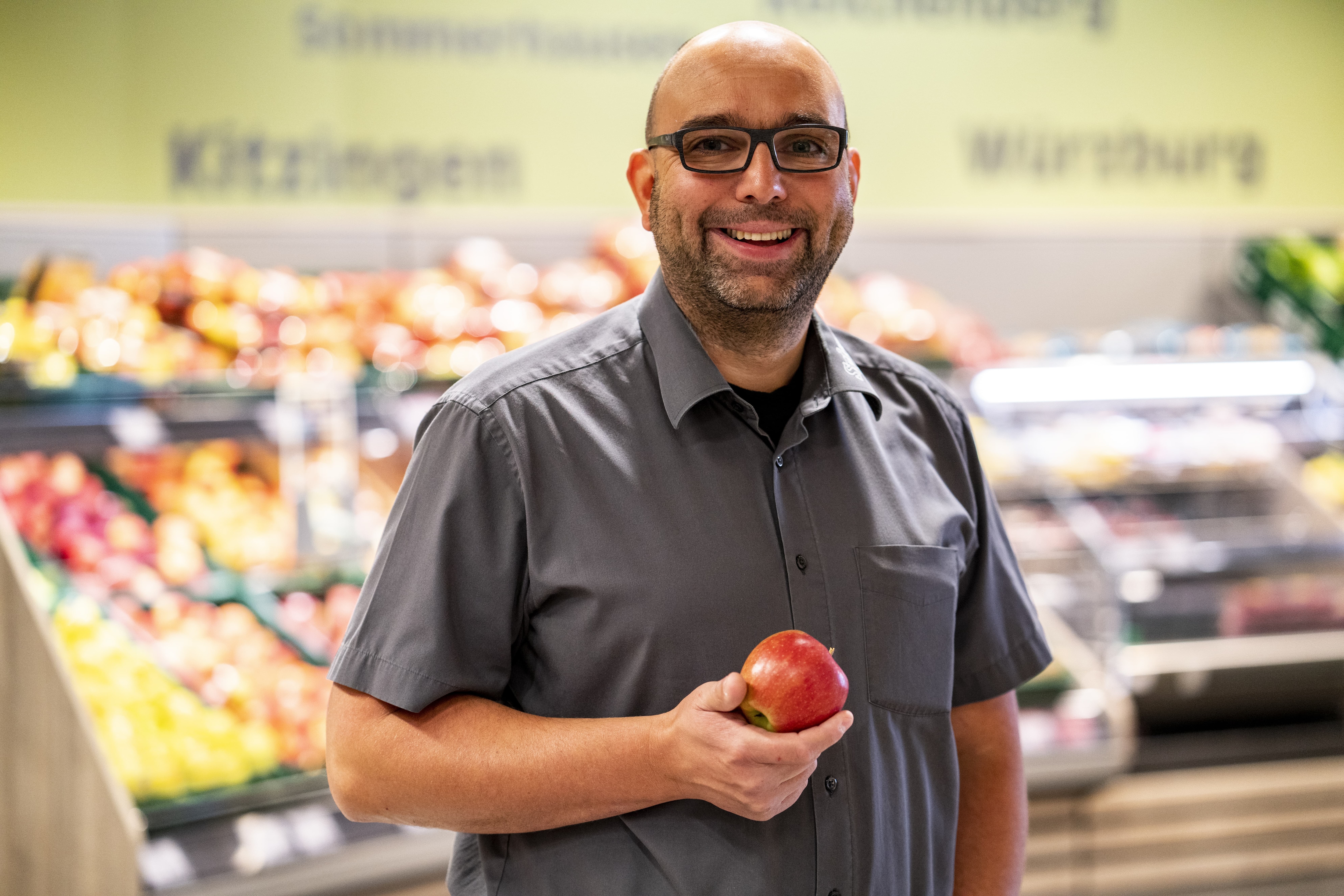 Der Marktleiter, lächelnd mit einem angebissenen Apfel in der Hand, im Hintergrund verschwommen die Obst- und Gemüseabteilung.
