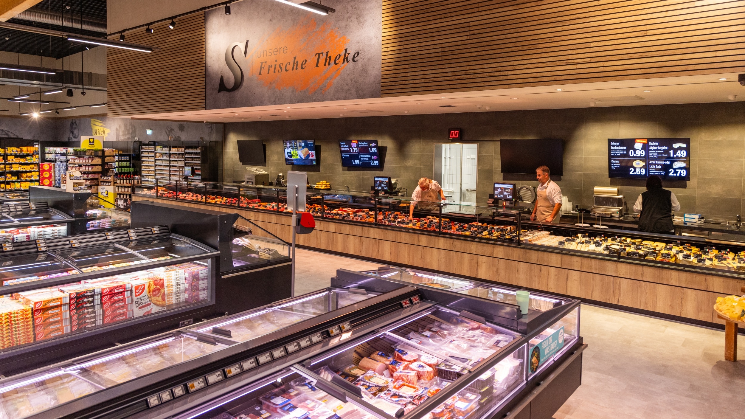 Gesamt-Front-Ansicht der Frische Theke mit drei Mitarbeiten. Im Vordergrund SB-Kühltruhen mit Produkten aus den Bereichen Wurst und Fleisch, Pizza und Fertiggerichte