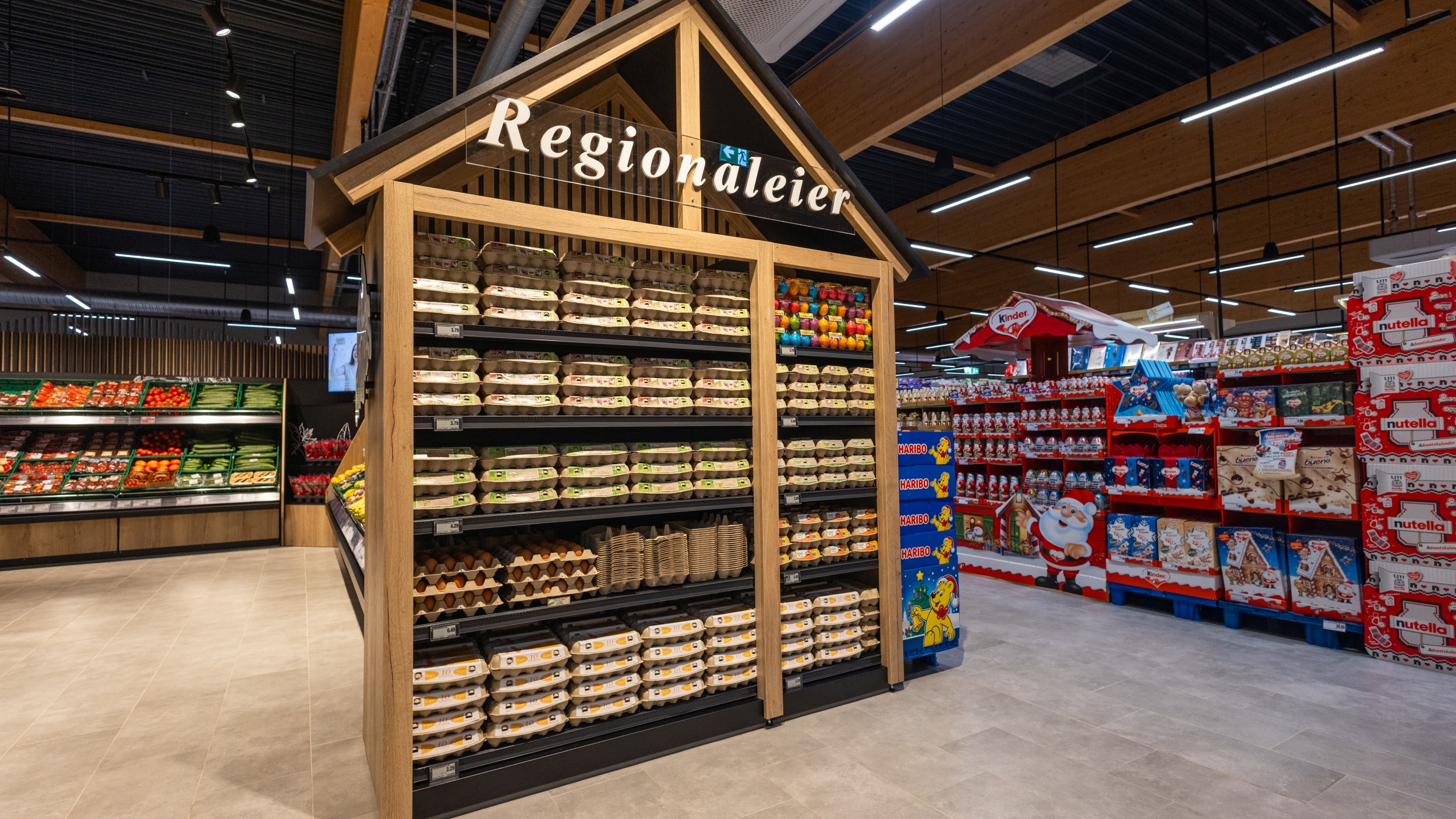 Im Bild befindet sich zentral ein Aufsteller in Form eines Hauses mit regionalen Eiern. Es werden sowohl lose als auch abgepackte Eier in unterschiedlichen Verpackungsgrößen angeboten, sowie bunte Eier. Im Gang rechts daneben werden Weihnachts-Süßigkeiten angeboten, links im Hintergrund ist ein Teil der Obst- und Gemüseabteilung zu erkennen