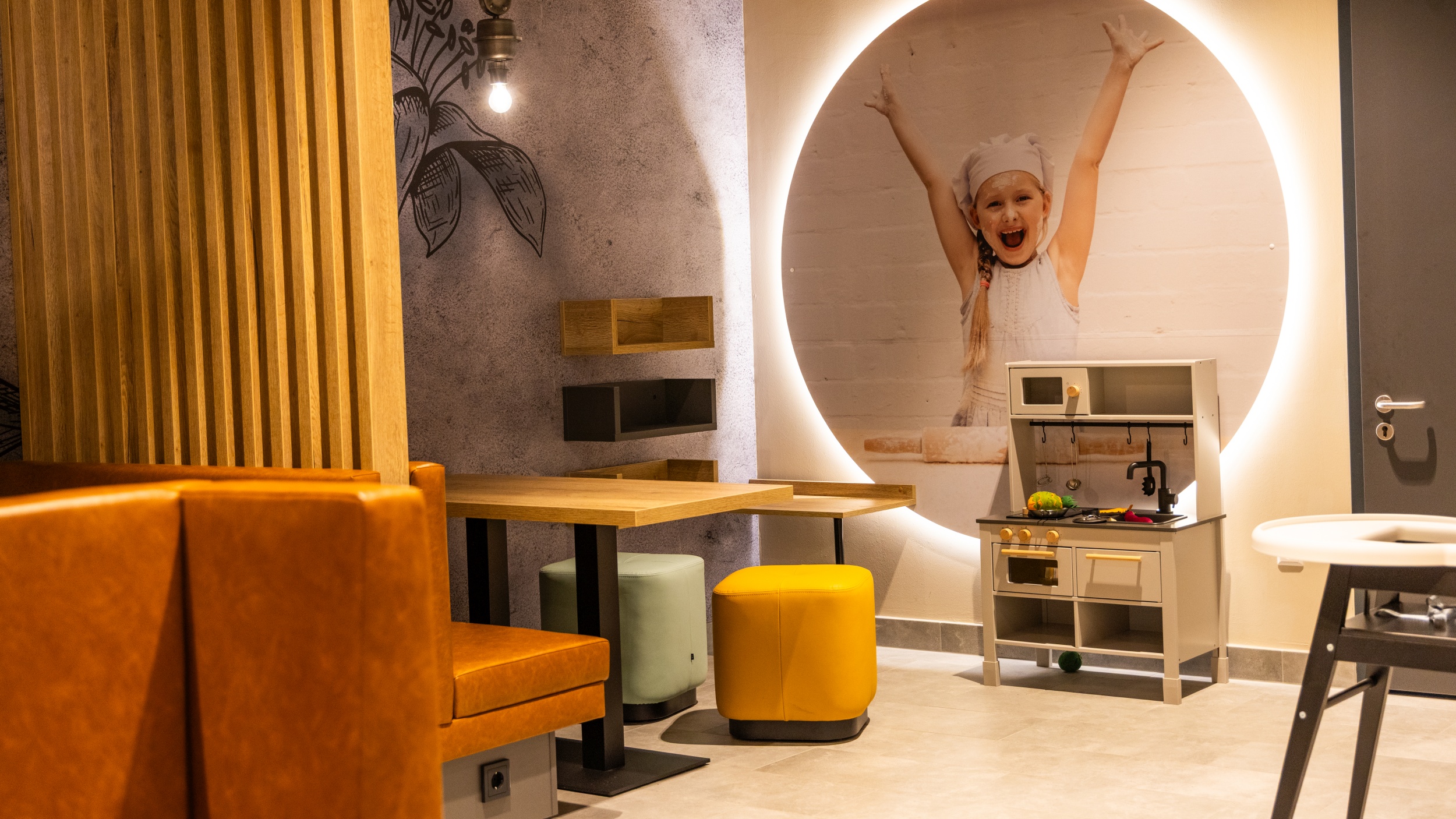 Spielecke für die Kleinen mit Kindersitzecke und Spielküche im Bereich des Restaurants. Im Hintergrund ein beleuchtetes Bild mit einem Mädchen mit Bäckermütze und Schürze