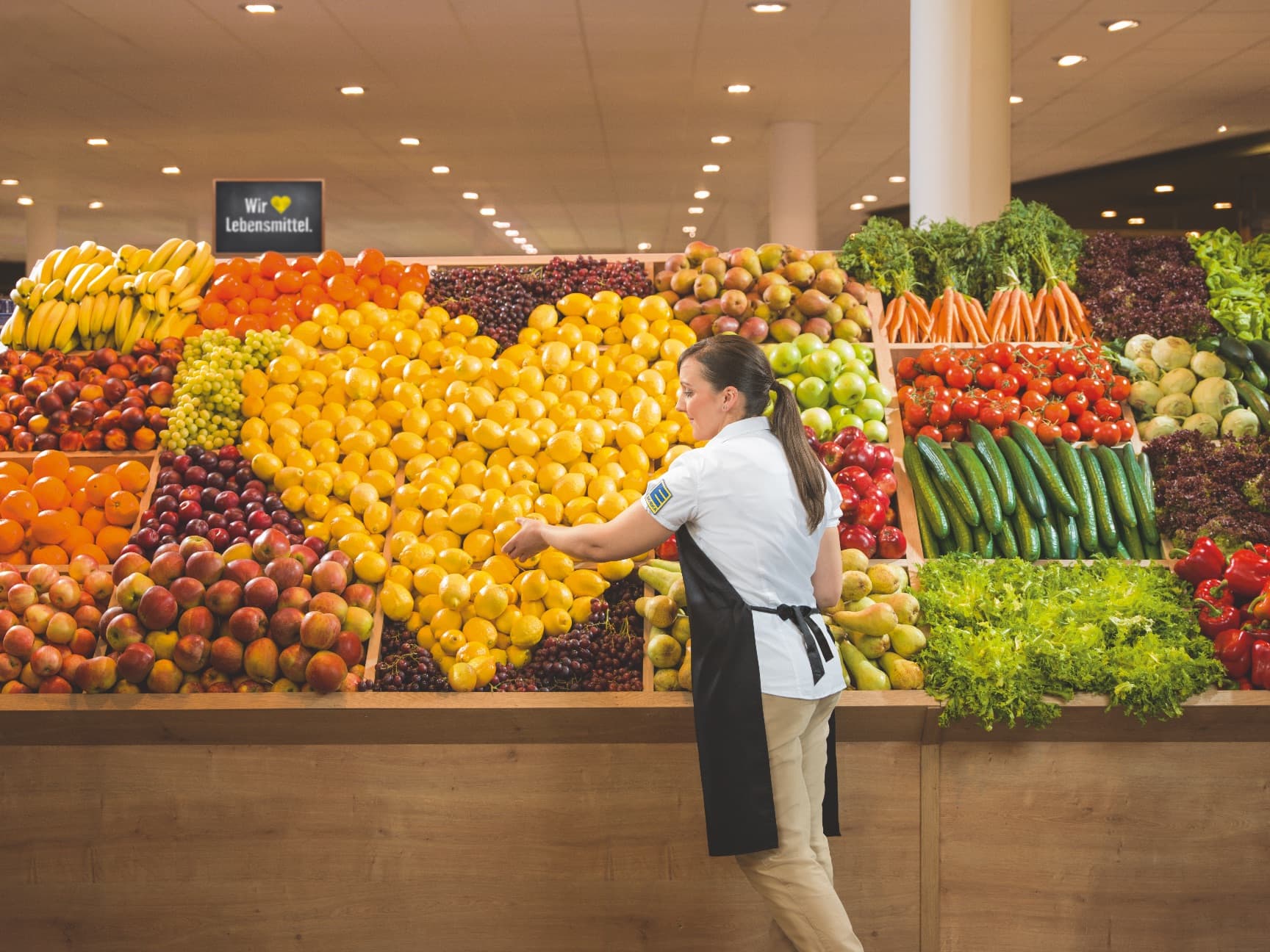 Eine junge Frau in EDEKA-Kleidung hat im Obstregal ein großes Herz aus gelben Zitronen gebastelt.