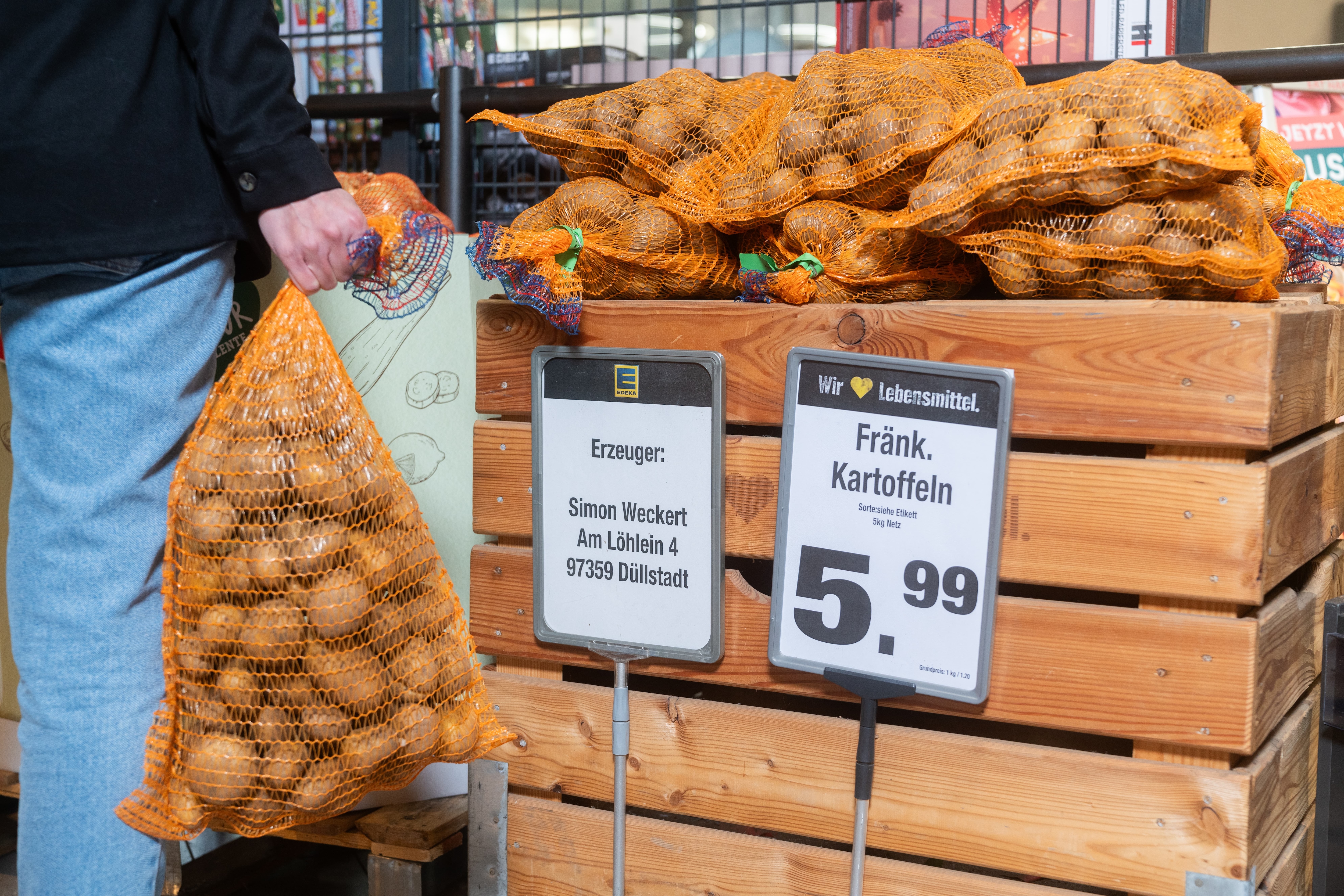 Eine Person steht mit einem Sack Kartoffeln in der Hand vor einer Holzkiste mit weiteren Kartoffelsäcken.