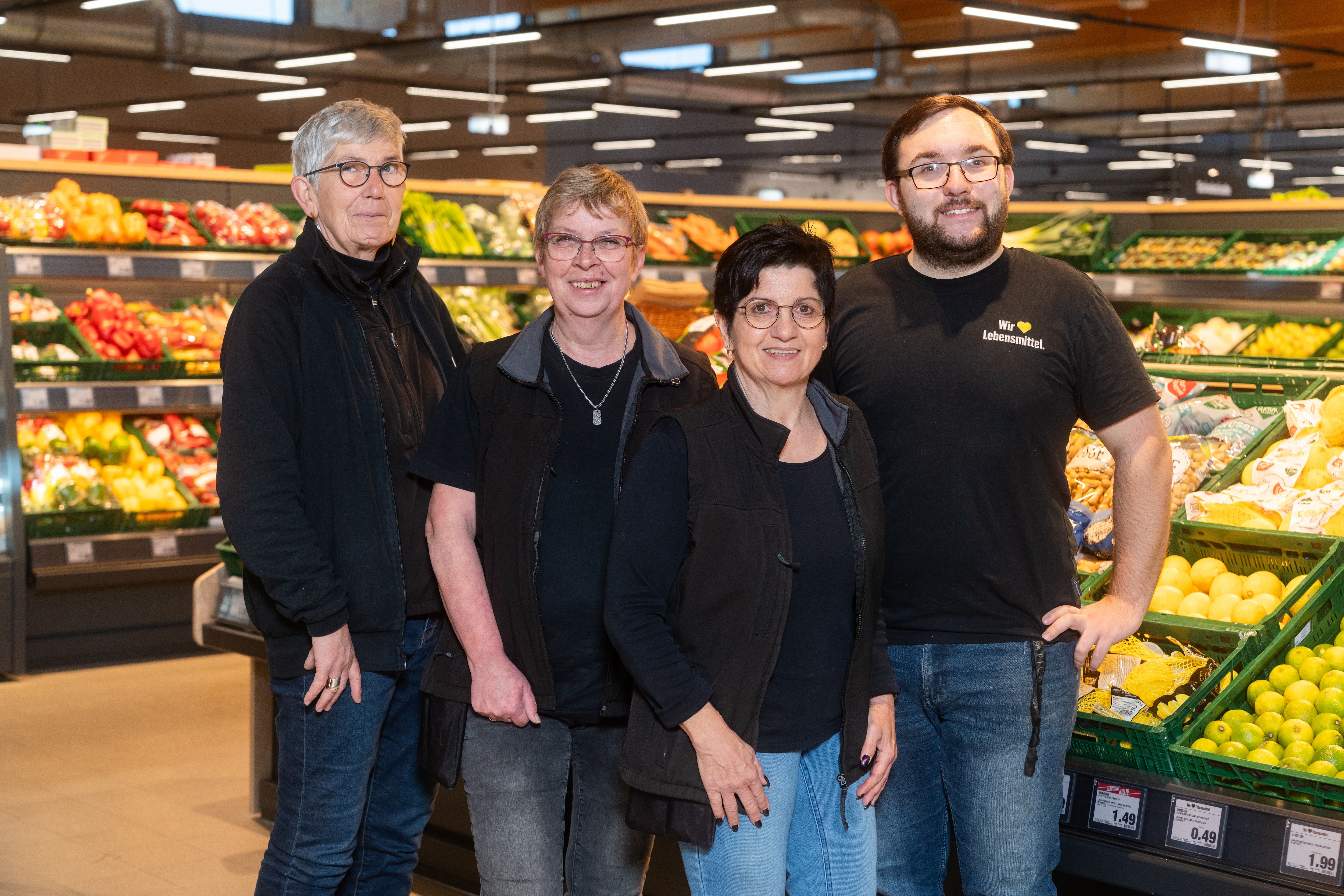 Drei Frauen und ein Mann in schwarzen Oberteilen stehen lächelnd in der Obstabteilung.