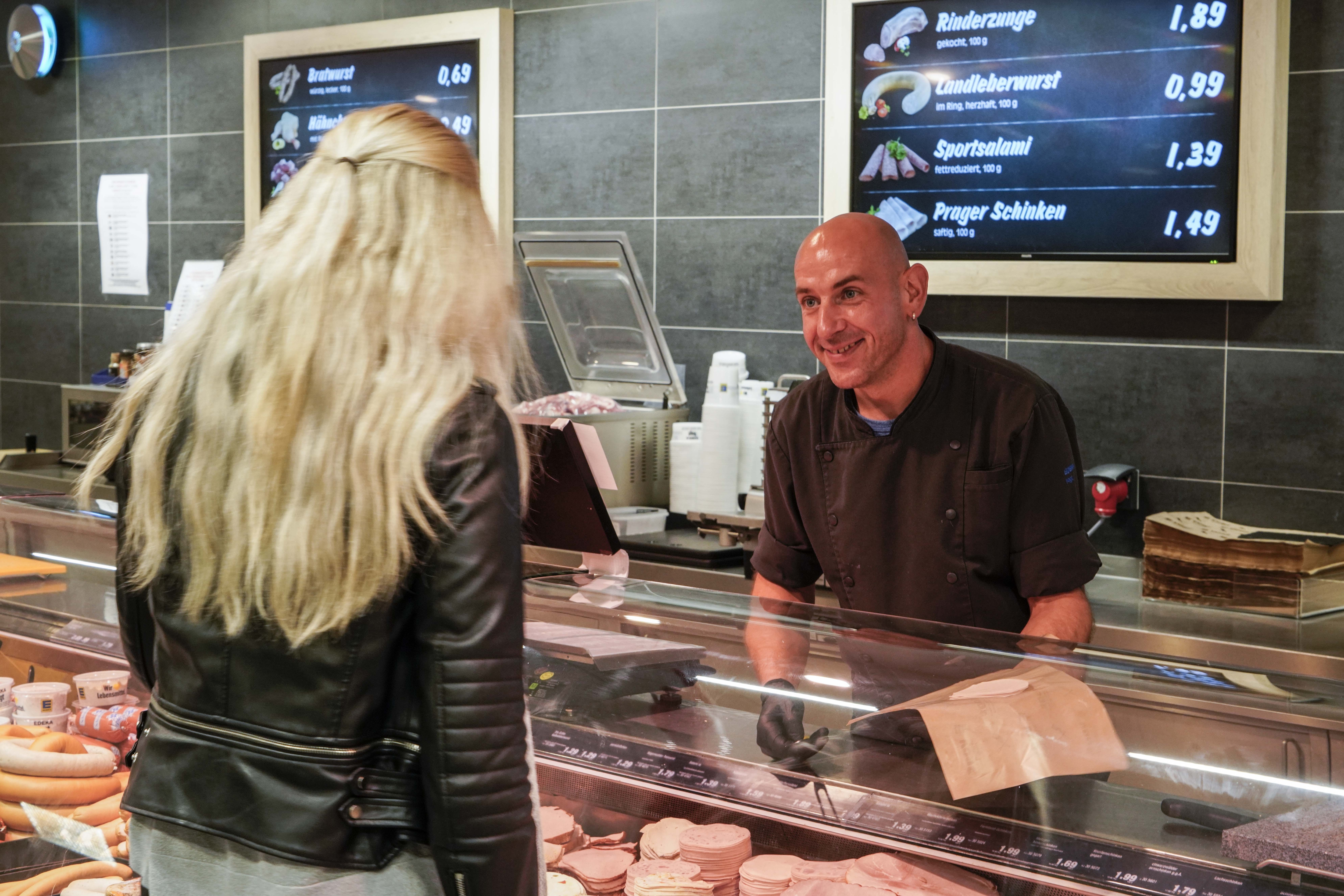 Ein Mitarbeiter in schwarzer Metzgerkluft unterhält sich über die Fleischtheke hinweg lächelnd mit einer Kundin.