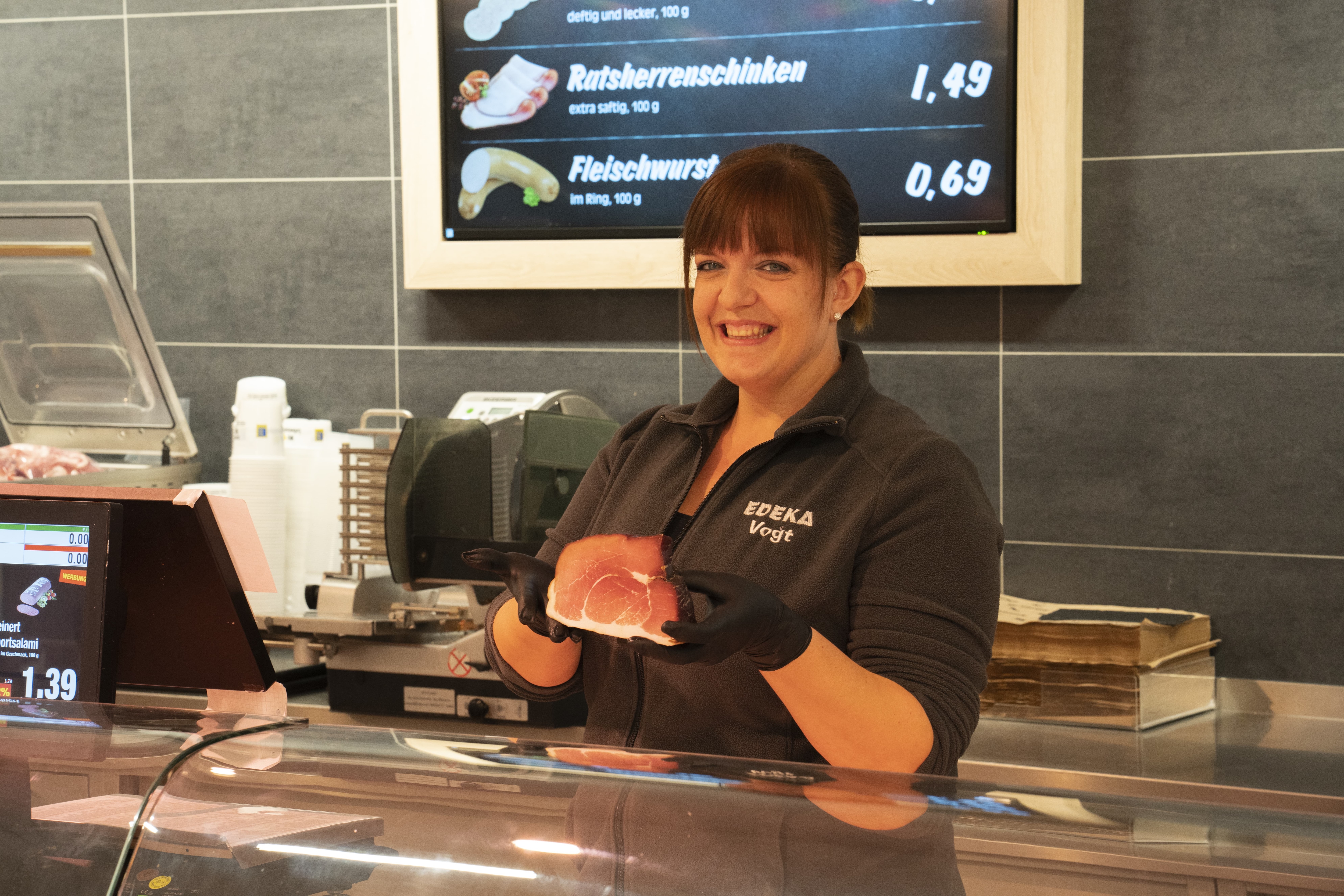 Eine junge Mitarbeiterin hinter der Bedientheke lächelt in die Kamera. Sie hält ein Stück Fleisch in den behandschuhten Händen.
