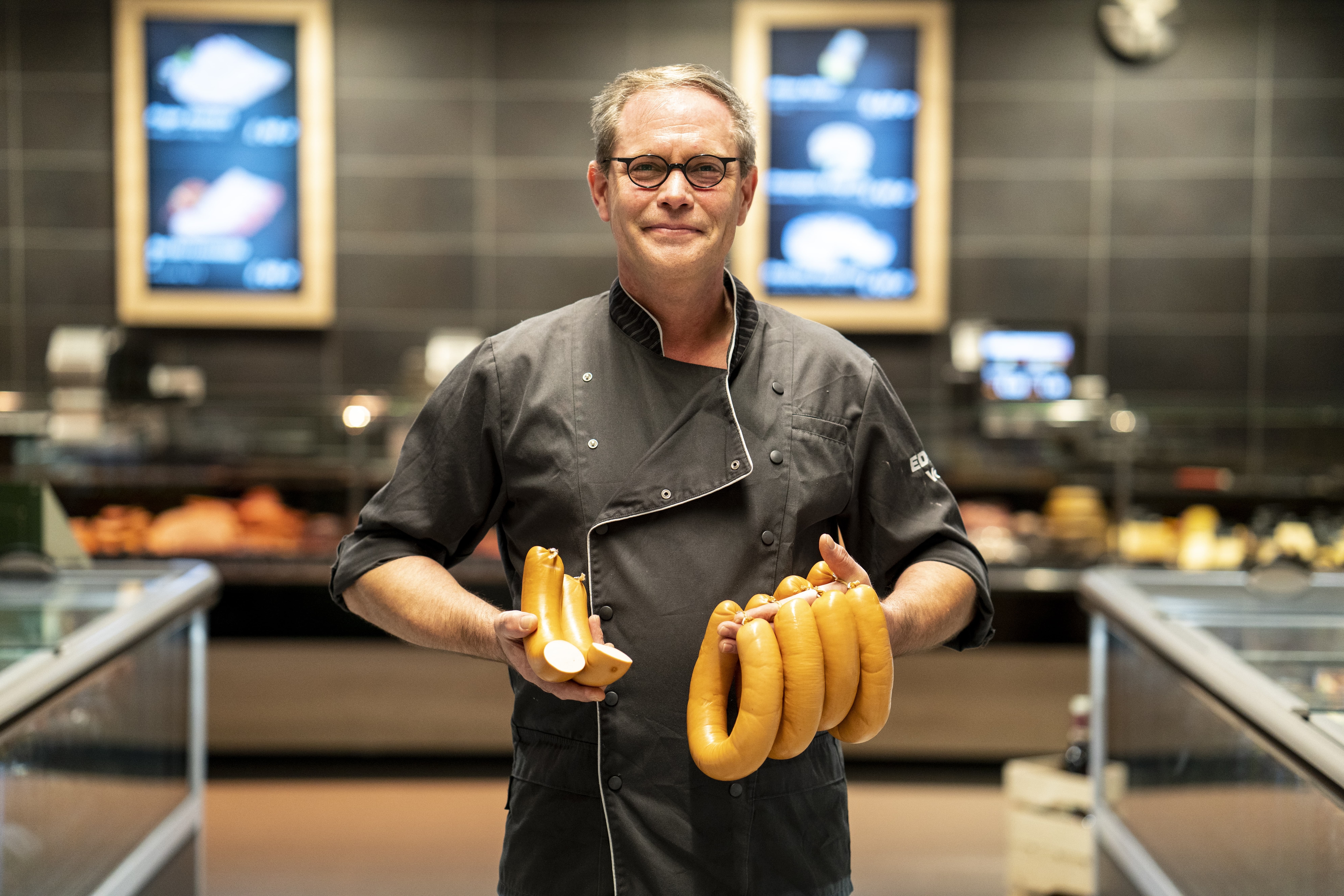 Ein Mitarbeiter in schwarzer Metzgerkluft lächelnd mit Wurst in den Händen zwischen den Tiefkühl-Gängen eines EDEKA-Markts.