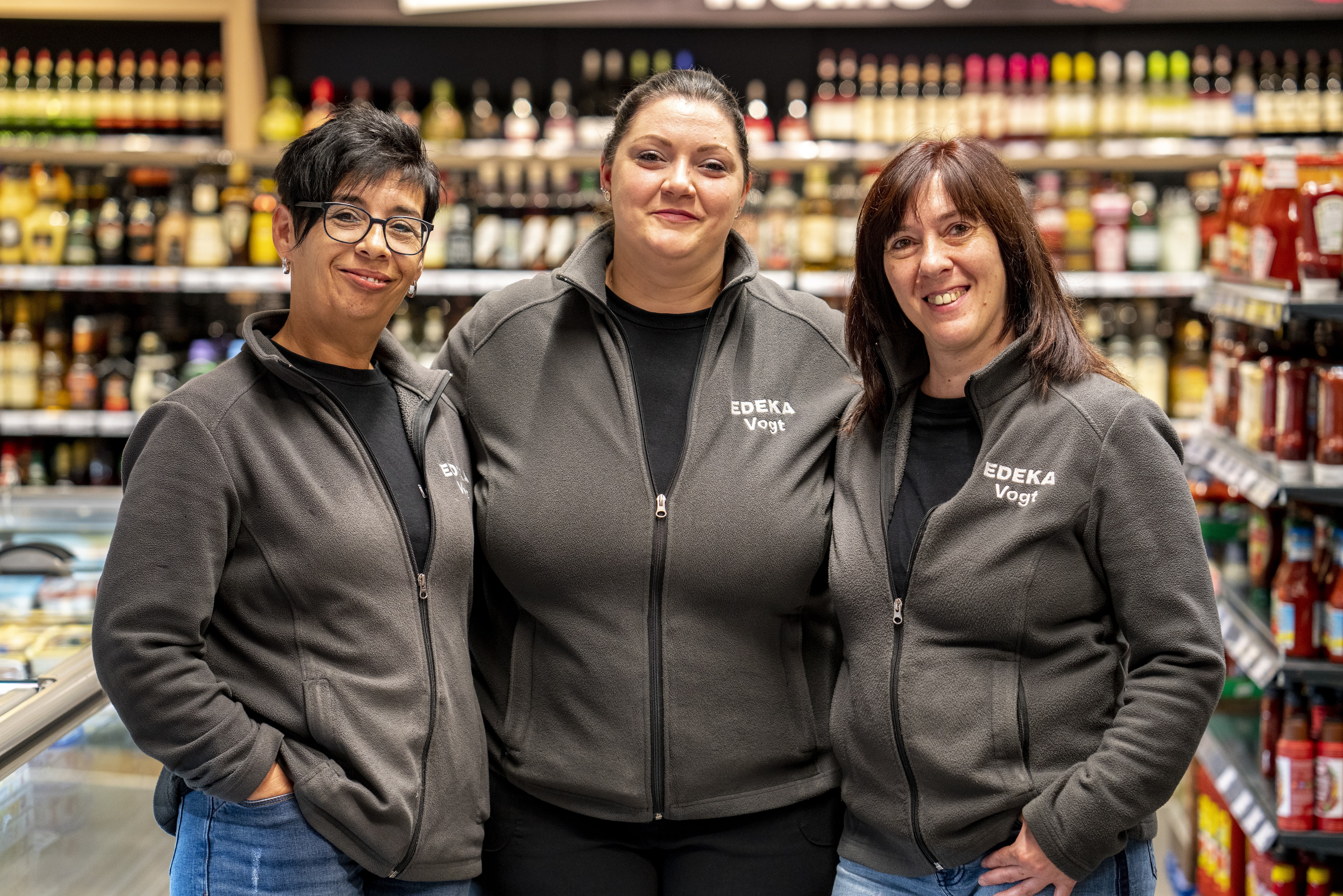 Drei Mitarbeiterinnen lächeln vor verschwommenen Produktregalen in die Kamera. Sie tragen graue Fleece-Westen, auf denen EDEKA Vogt steht.