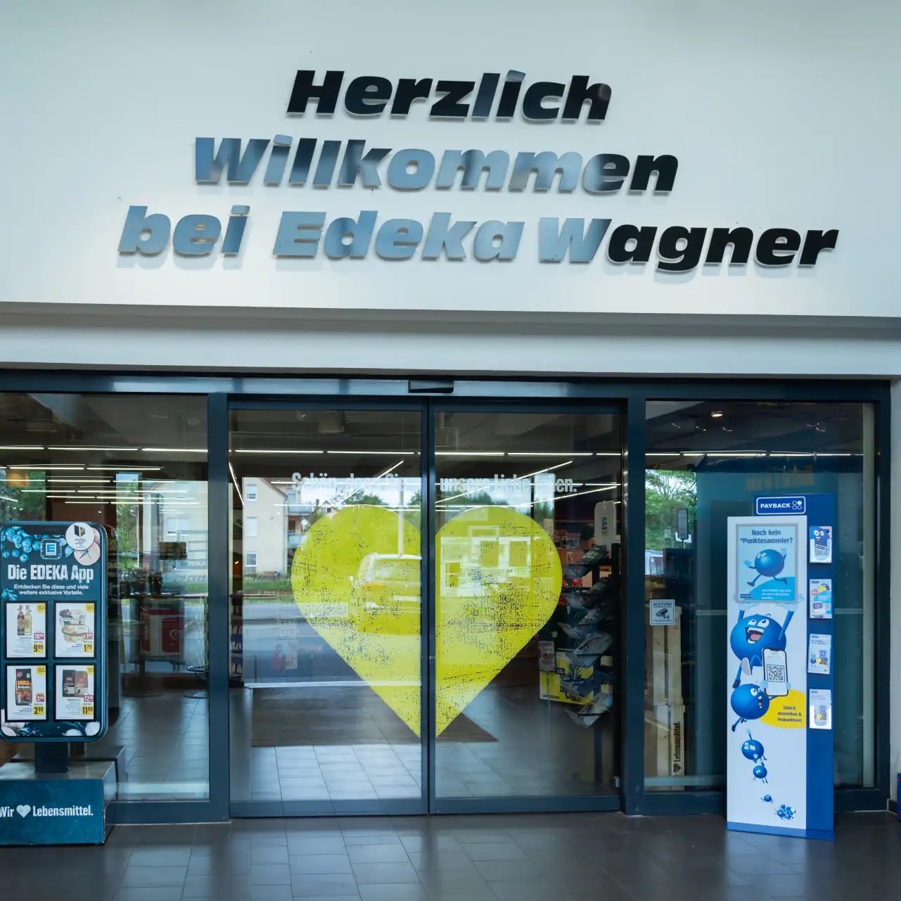 Eingangstür von Edeka Wagner mit einem großen gelben Herz auf der Schiebetür und oberhalb an der Wand ein Schriftzug mit Herzlich Willkommen bei Edeka Wagner.