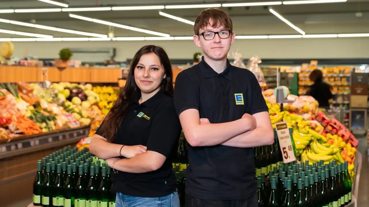 Zwei Azubis stehen in der Obstabteilung mit verschränkten Händen und Rücken an Rücken.