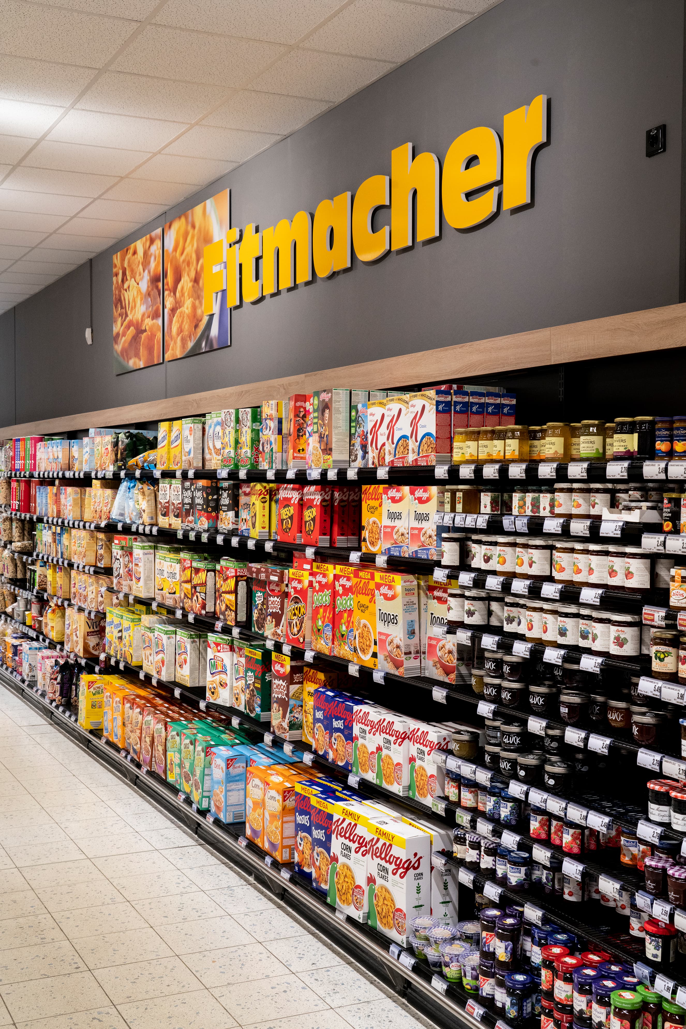 Ein Regal mit Frühstückscerealien und Marmelade, über dem an der Wand steht: Fitmacher.