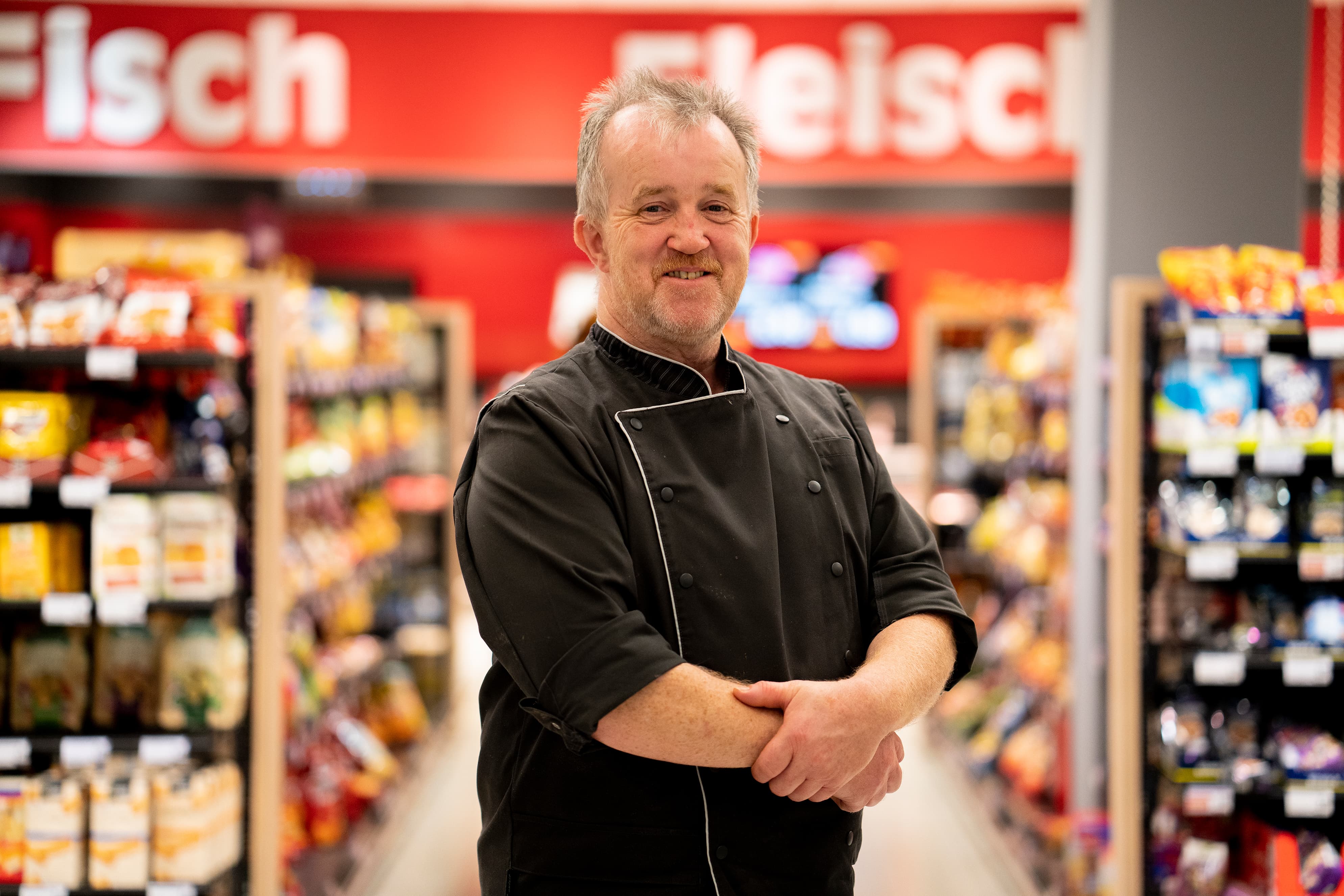 Ein Mitarbeiter in schwarzer Metzgerkluft lächelnd zwischen den Gängen eines EDEKA-Markts.