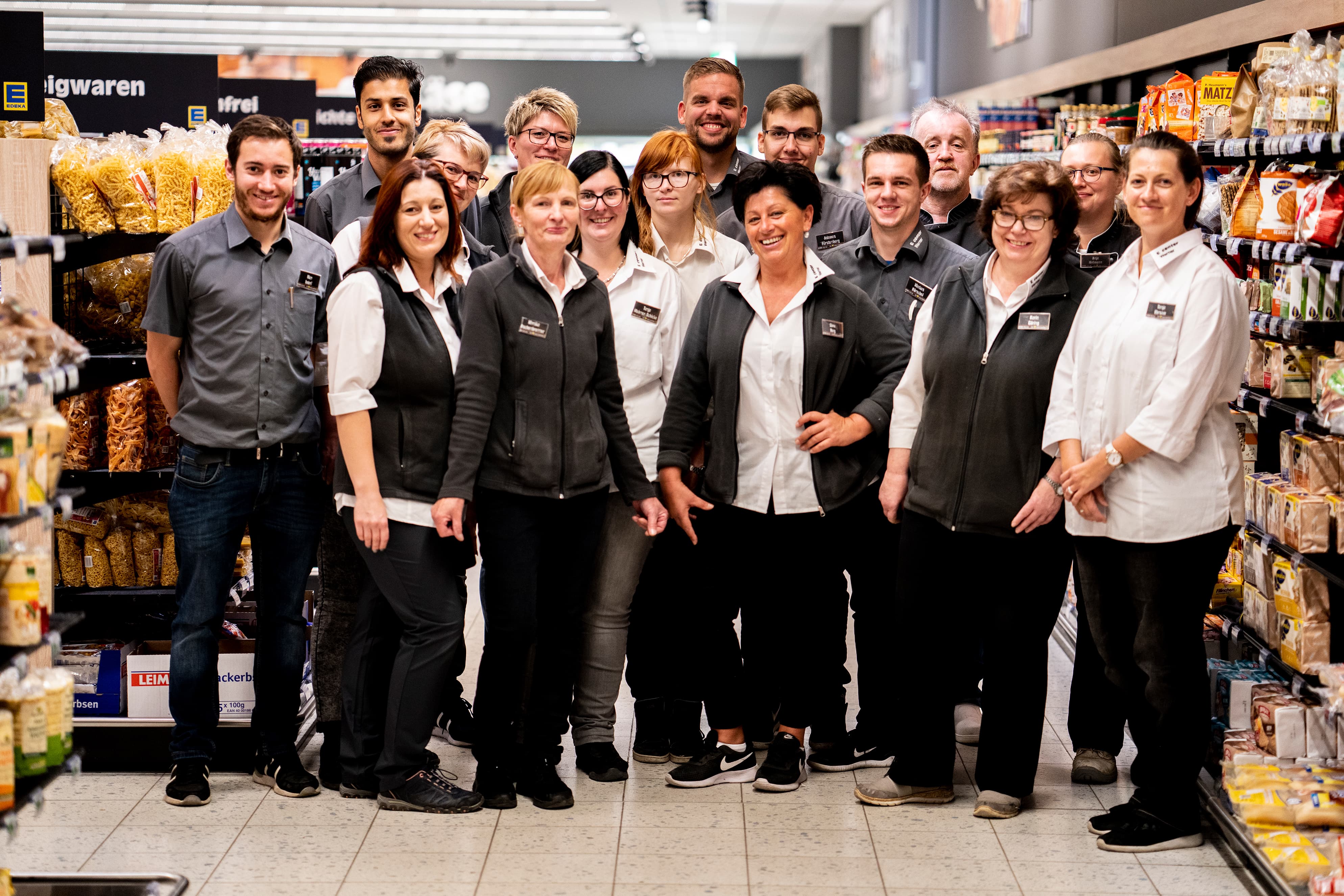 Gruppenfoto des Teams dieses EDEKA-Markts, aufgenommen im Gang des Markts zwischen Nudeln und Knäckebrot. Alle lächeln.