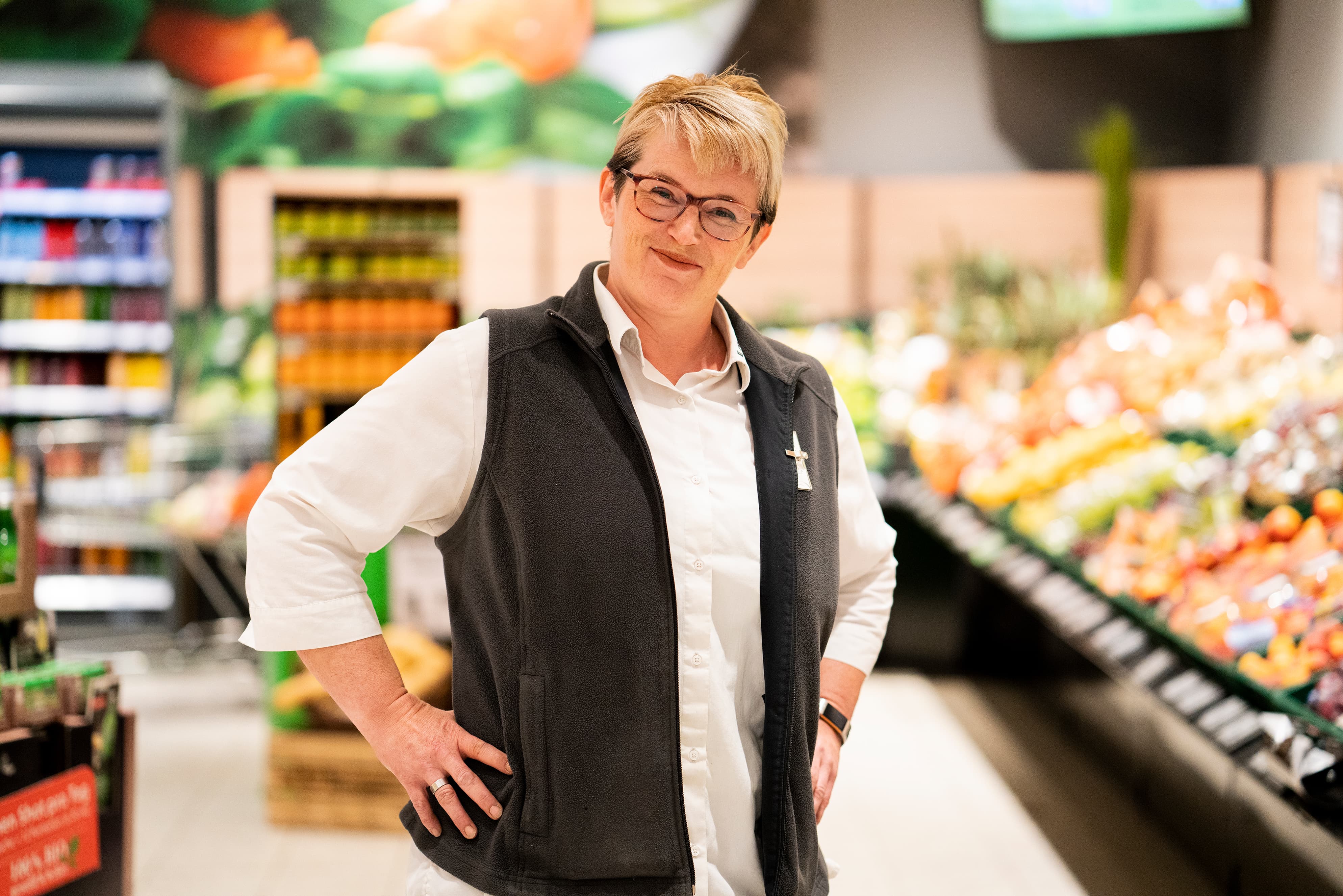 Eine Mitarbeiterin mit blonden Haaren steht lächelnd in der Gemüseabteilung eines EDEKA-Markts.