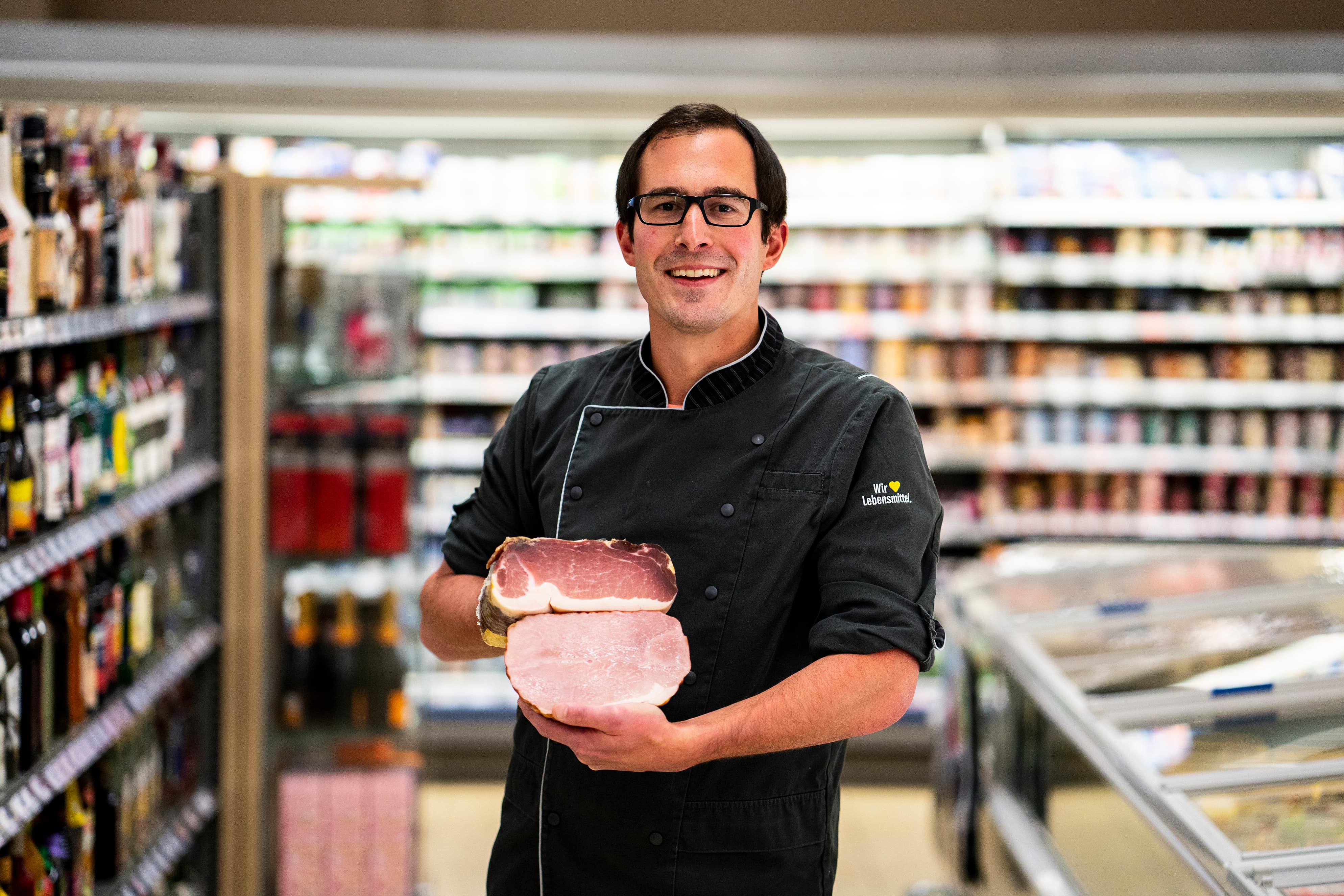 Ein lächelnder Mitarbeiter in der schwarzen Kluft eines Metzgers steht im Gang eines EDEKA-Markts und hält ein Stück Fleisch in beiden Händen.