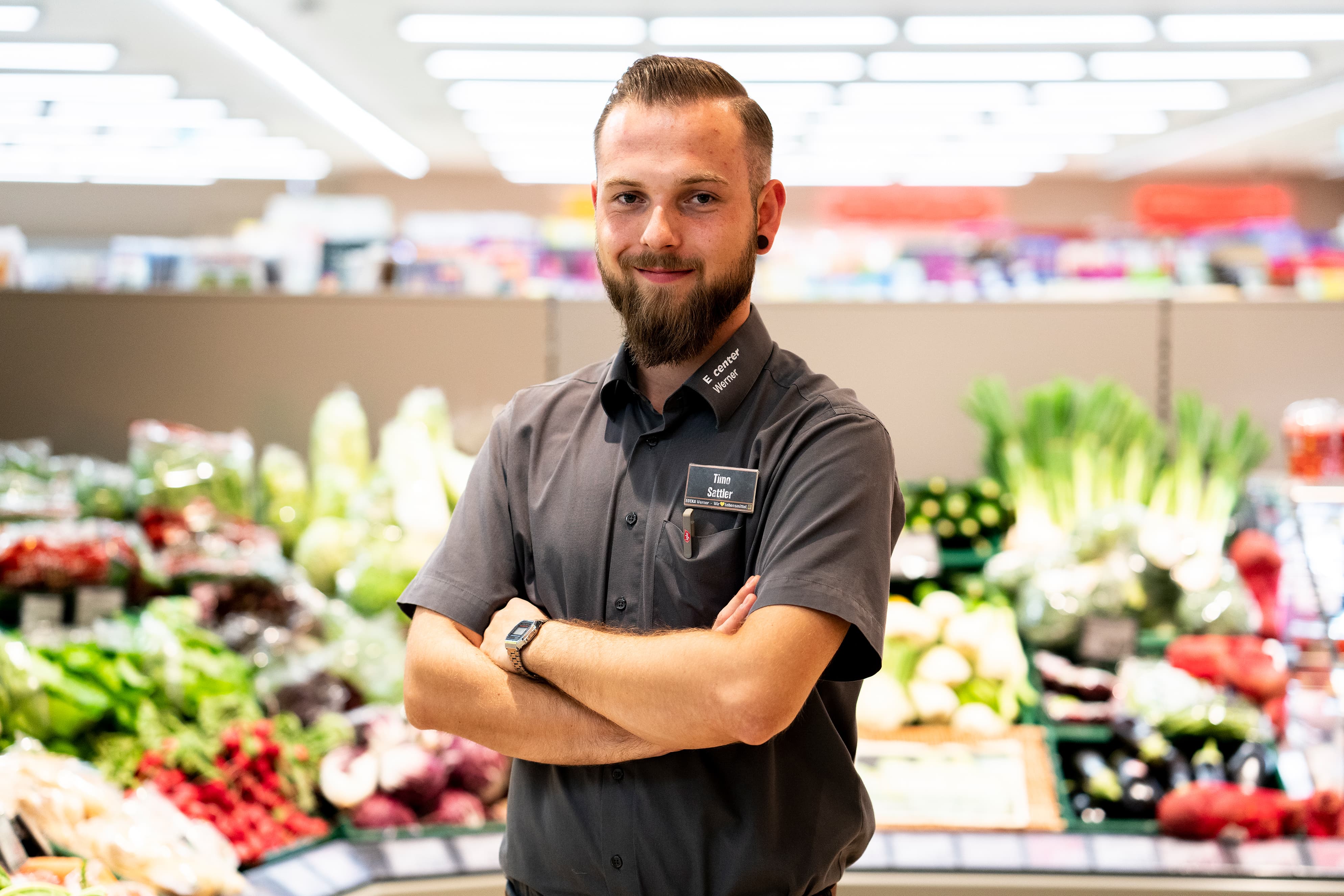 Ein junger Mann mit Bart und verschränkten Armen steht vor der Gemüseabteilung eines EDKA-Markts.