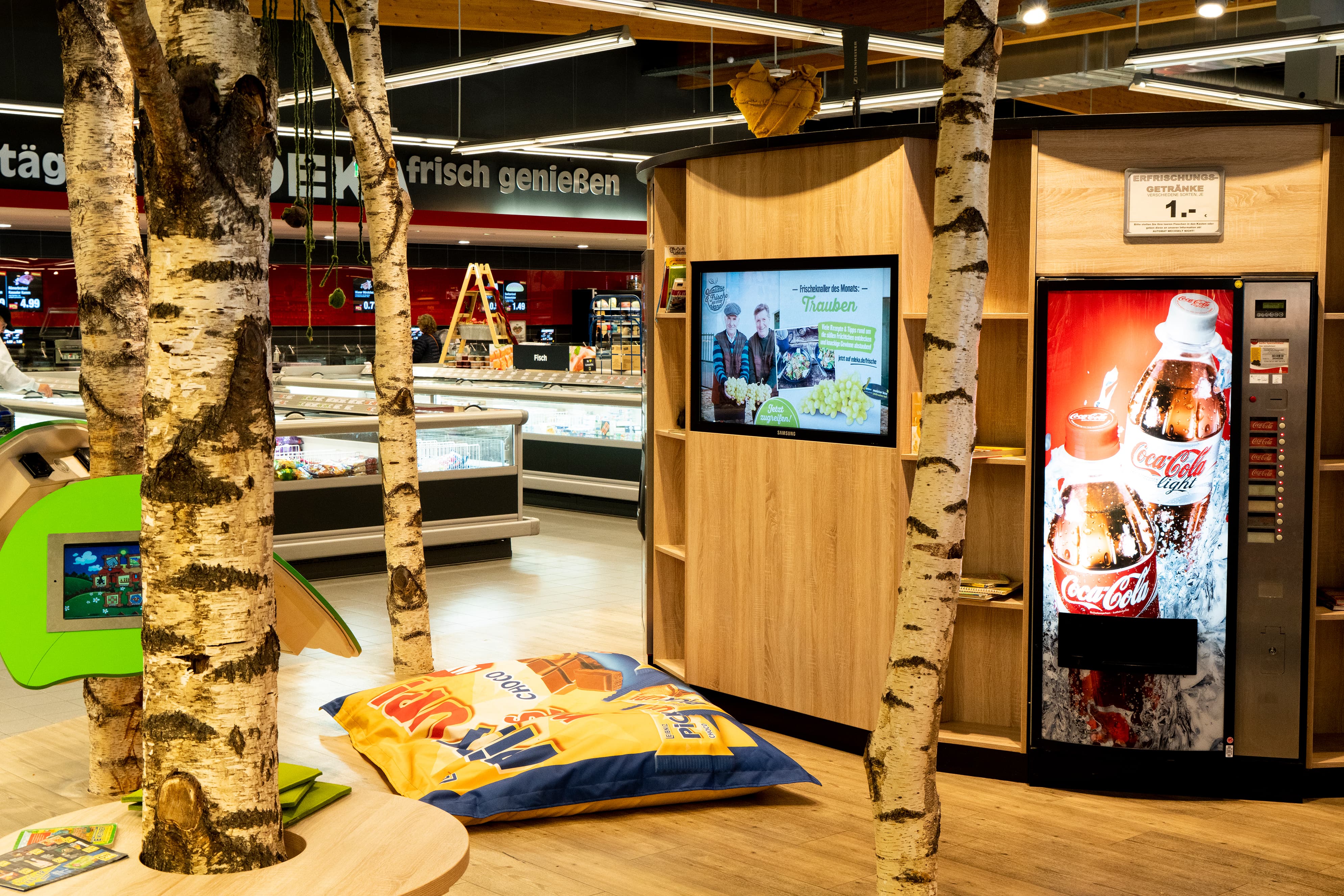 Ein Bereich in einem EDEKA-Markt, der zum Aufenthalt einlädt. Es gibt einen Fernseher, einen Getränkeautomaten, ein Sitzkissen am Boden und mehrere Birkenstämme zur Dekoration.