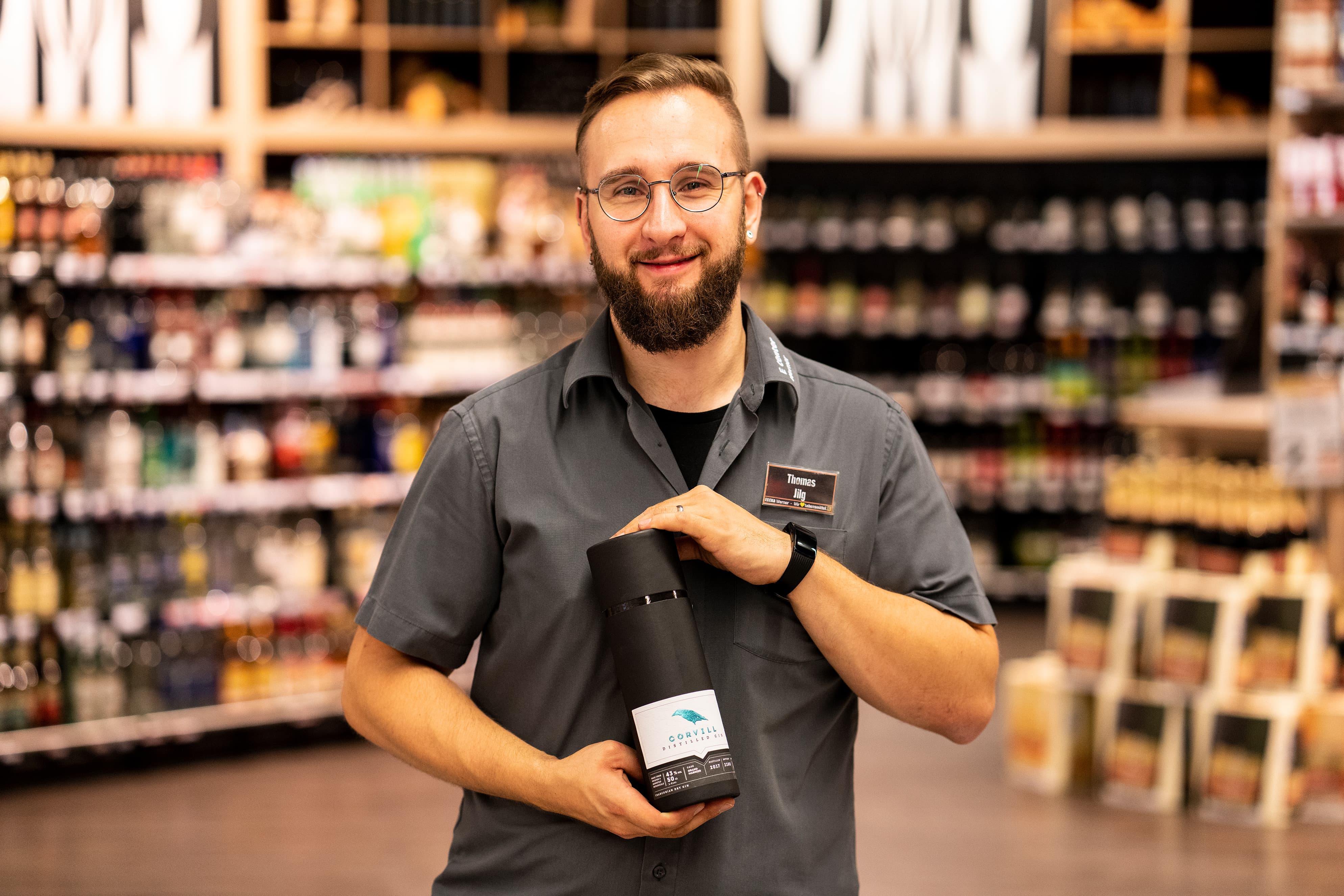 Ein Mitarbeiter steht lächelnd in der Alkoholabteilung eines EDEKA-Markts. Er hält eine Gin-Flasche in den Händen, die aussieht wie eine Thermoskanne.