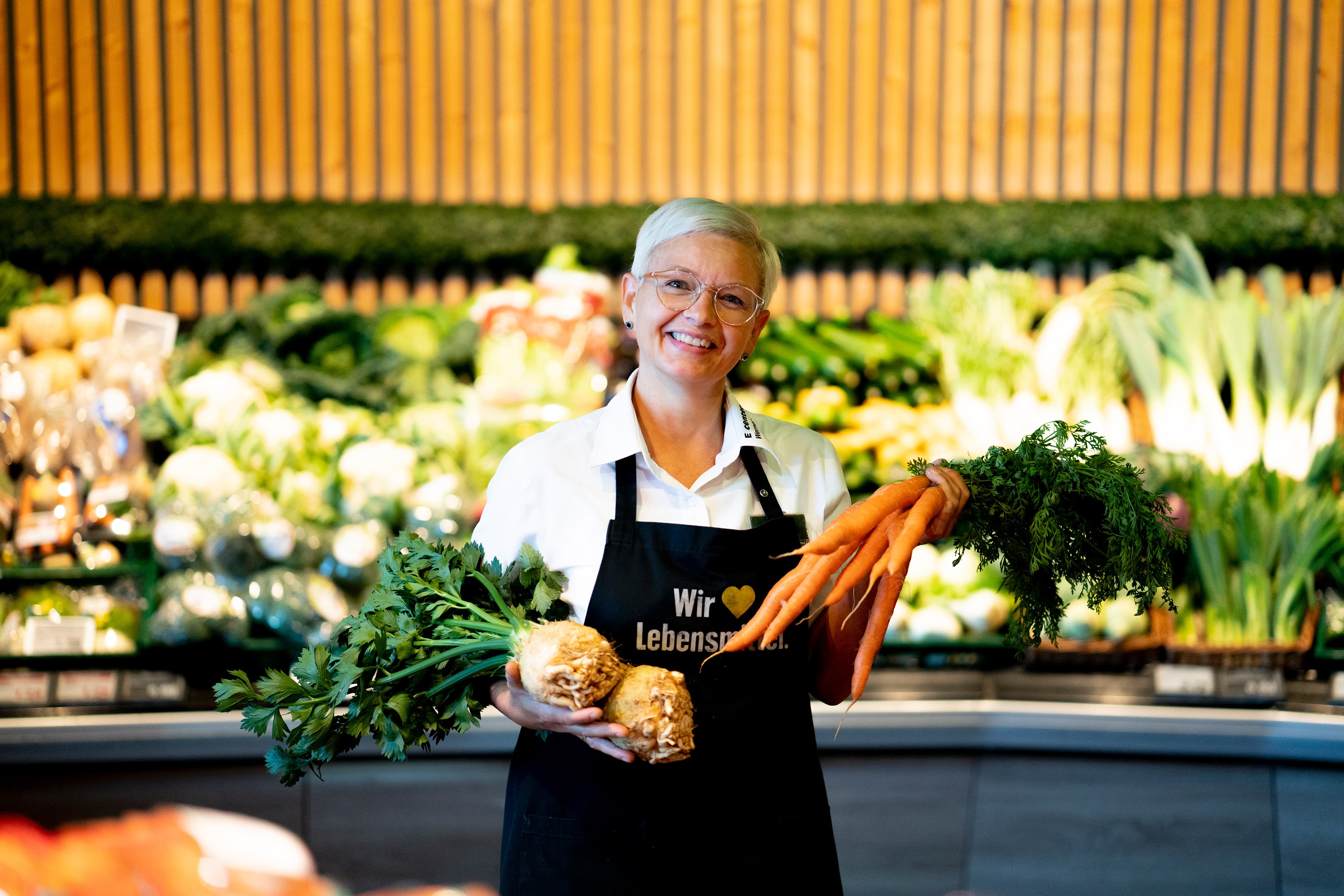 Eine Mitarbeiterin mit blonden Haaren steht lächelnd in der Gemüseabteilung eines EDKA-Markts.