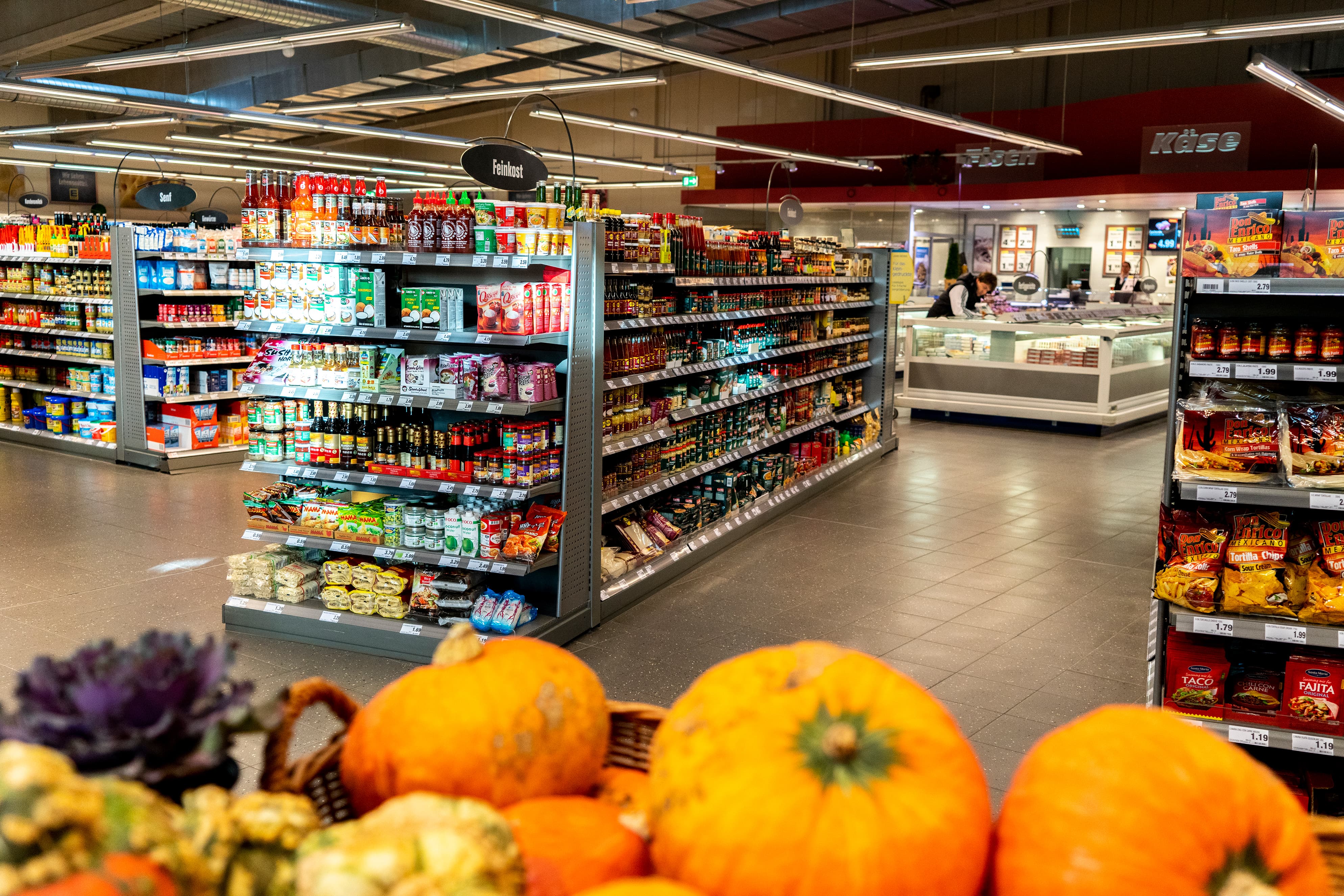 Gänge voller Produkte reihen sich in einem EDEKA-Markt auf. Im Vordergrund Kürbisse.