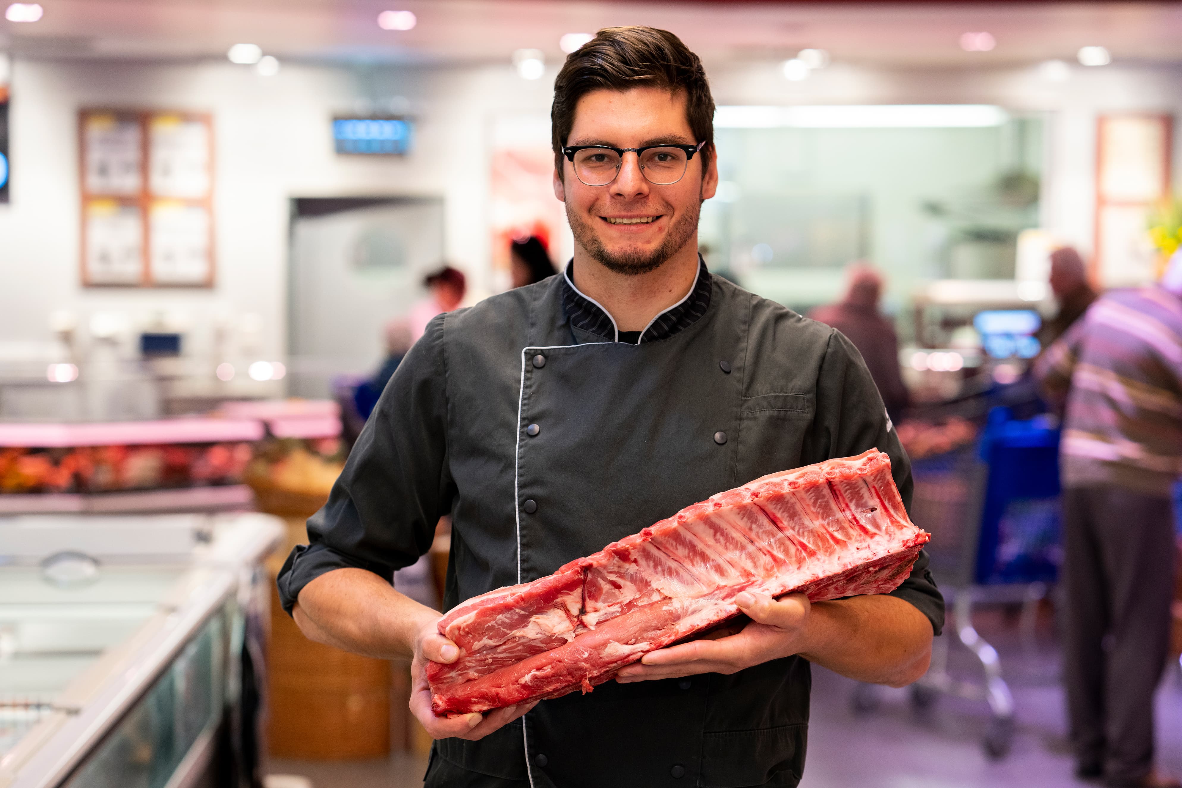 Ein junger Mitarbeiter in schwarzer Metzgerkluft lächelnd zwischen den Gängen eines EDEKA-Markts und hält ein großes Stück Fleisch in den Armen.