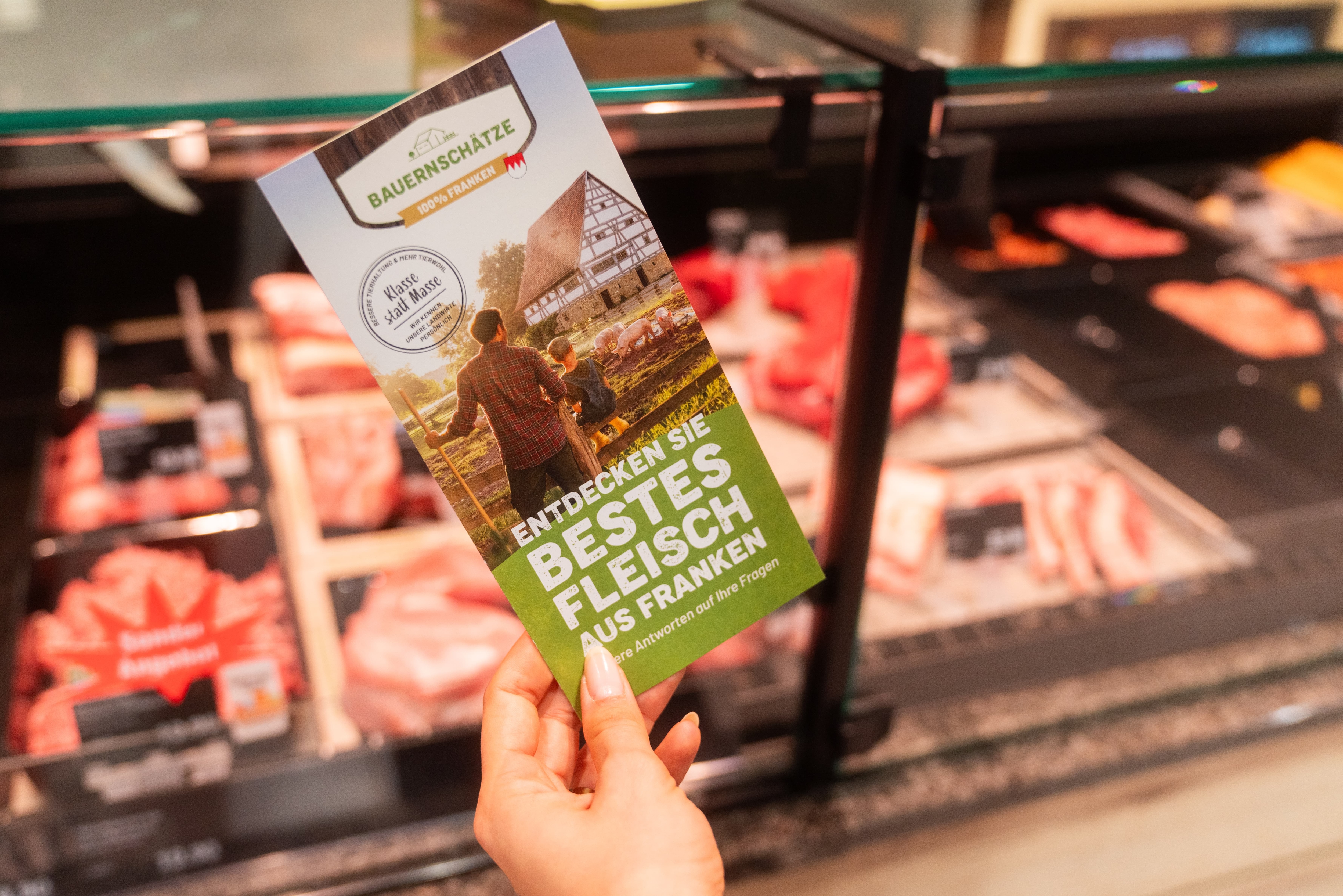 Eine Hand hält einen Flyer vor der Frischetheke. Auf ihm steht: Entdecken Sie bestes Fleisch aus Franken.