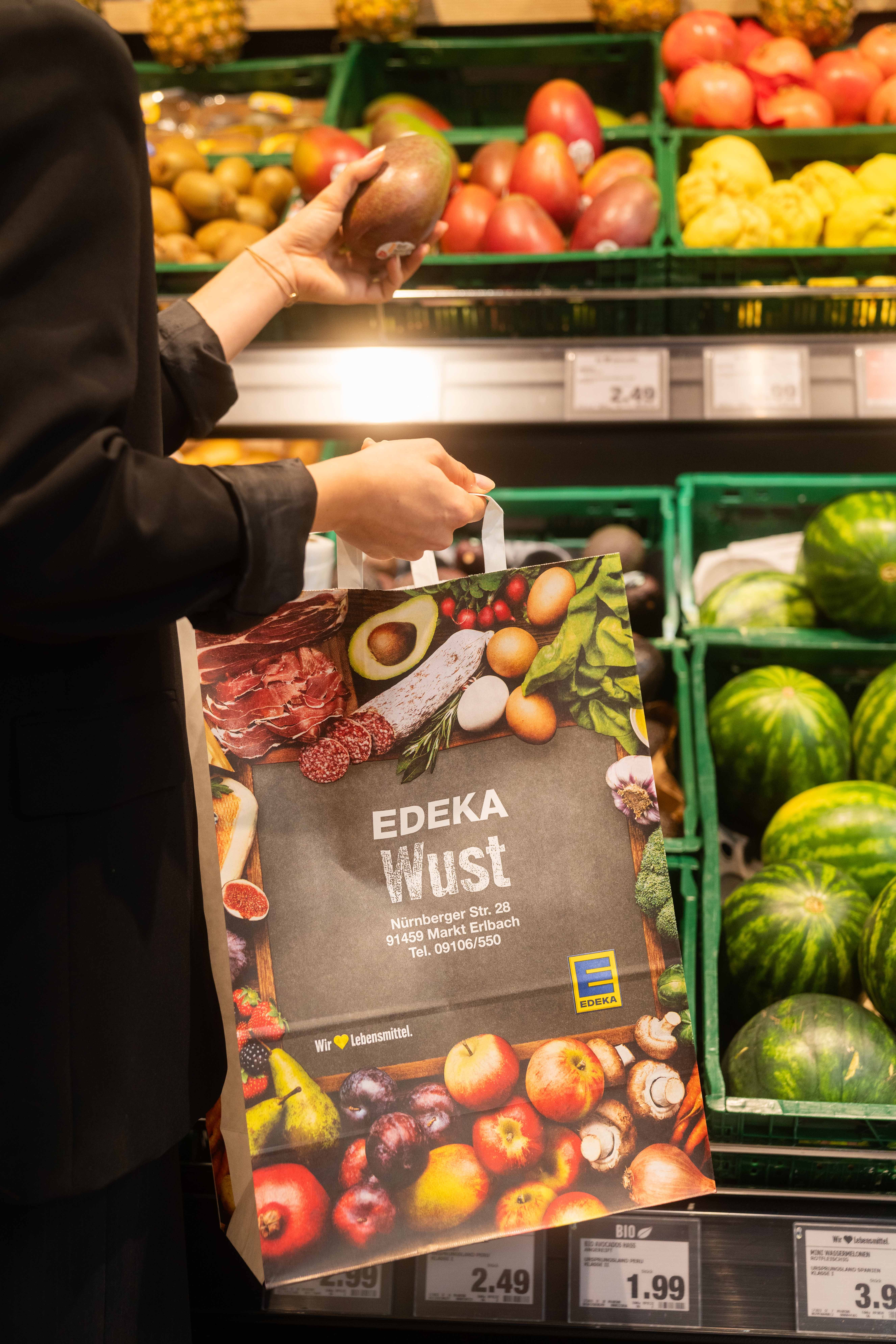 Eine Person steh am Obstregal und hat in der einen Hand eine Mango und in der anderen eine Papiertüte mit dem Schriftzug EDEKA Wust darauf.