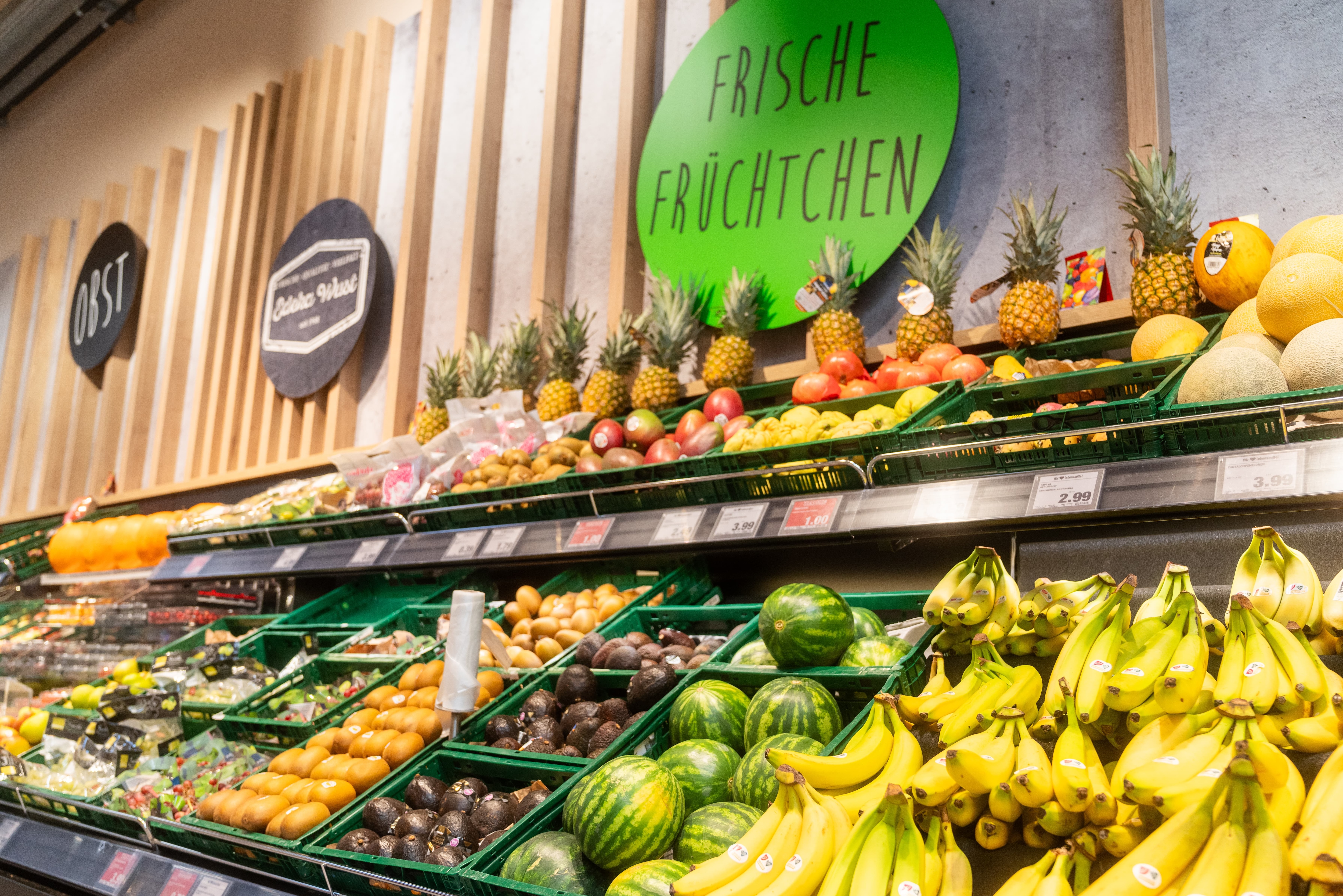 Über dem Obstregal mit Bananen, Melonen, Avocados und Kiwis steht „Frische Früchtchen.“