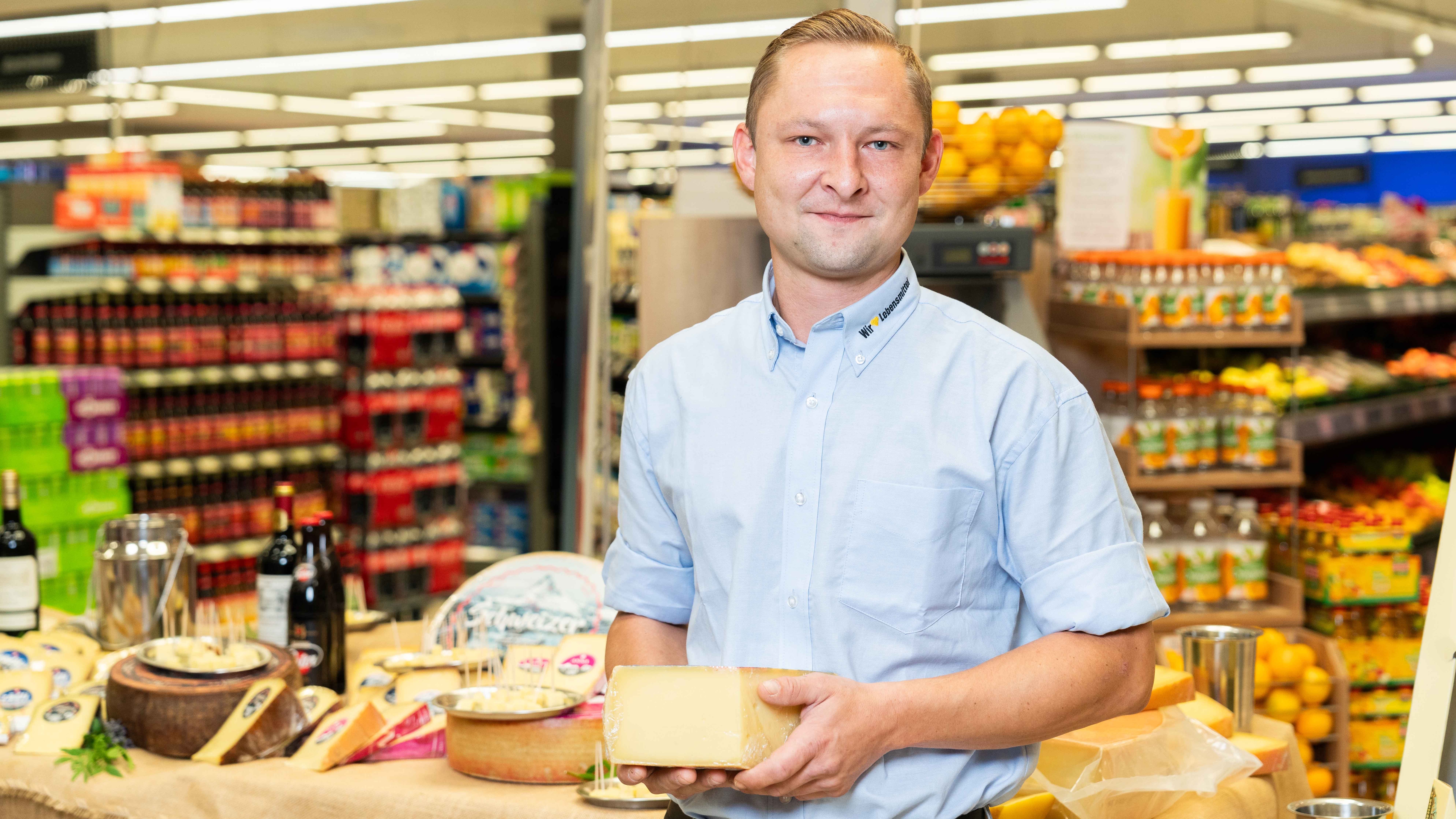 Der Käsesommellier steht vor der Auslage mit Käse und hält ein Stück Käse in der Hand.