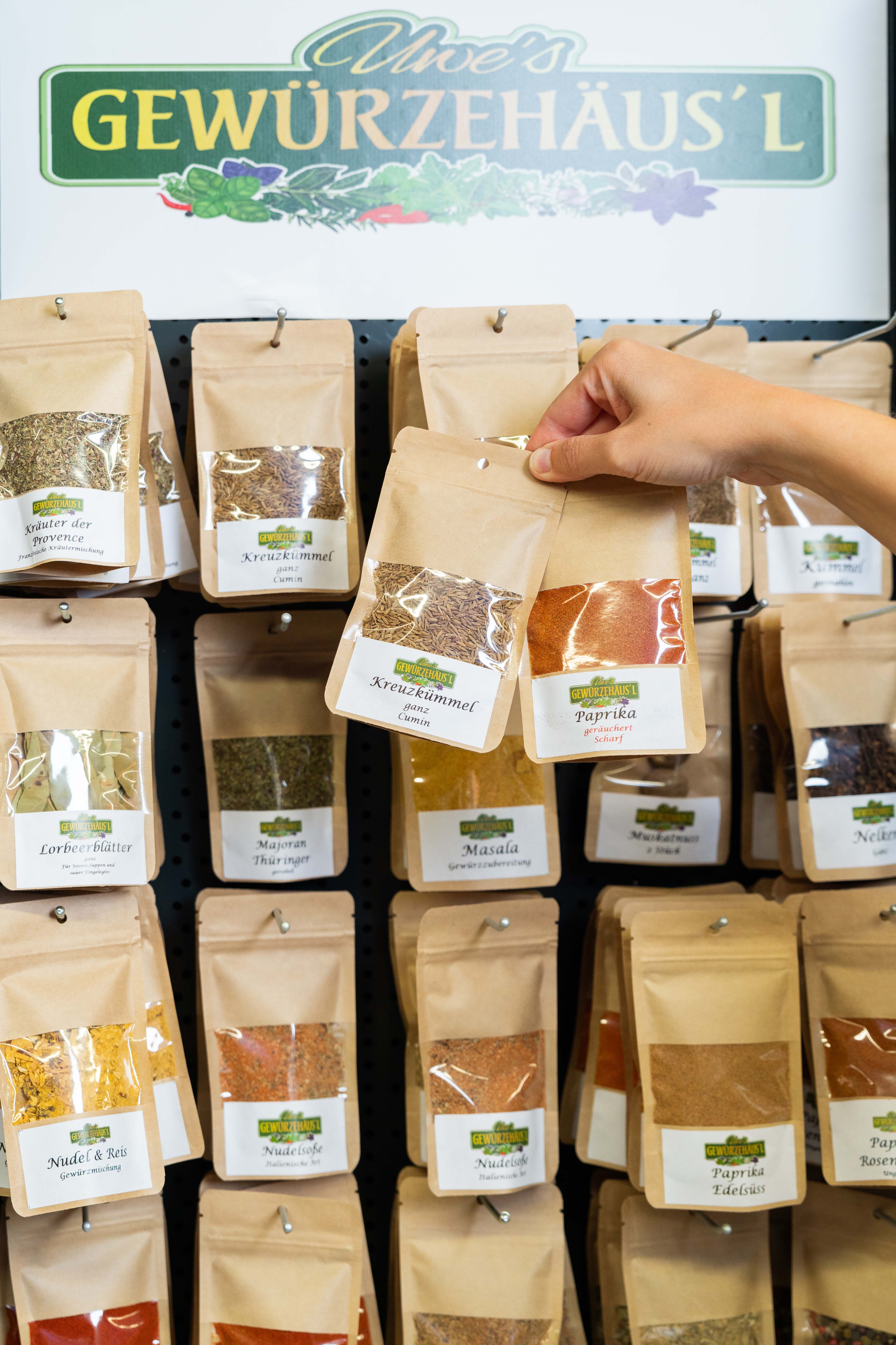 Aufsteller von Uwe's Gewürzhäusl mit verschiedenen Gewürzpackungen. Zwei davon werden von einer Hand davor gehalten.