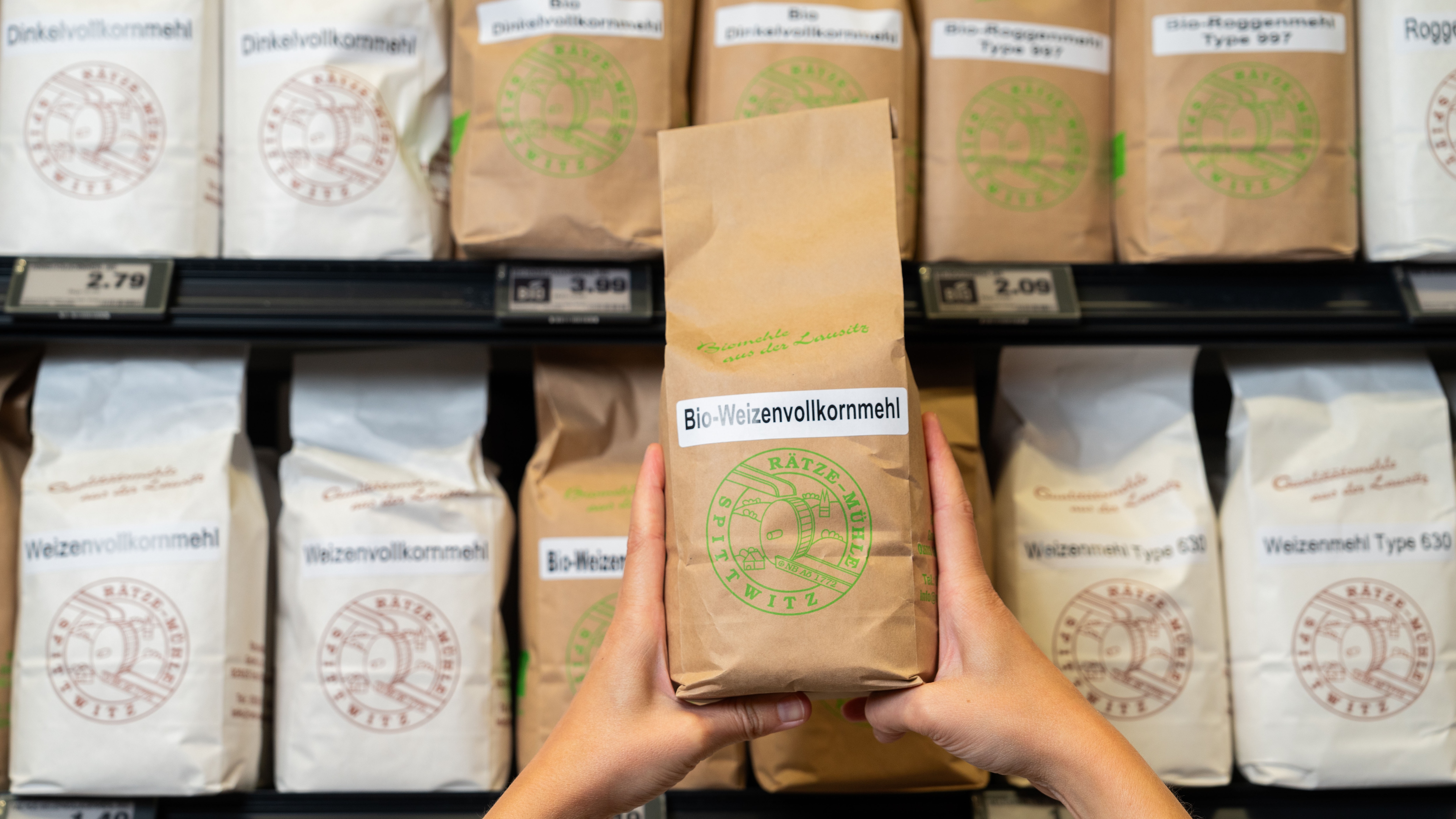 Vor dem Mehr-Regal wird eine Packung Mehl der Rätze Mühle mit beiden Händen hochgehalten.