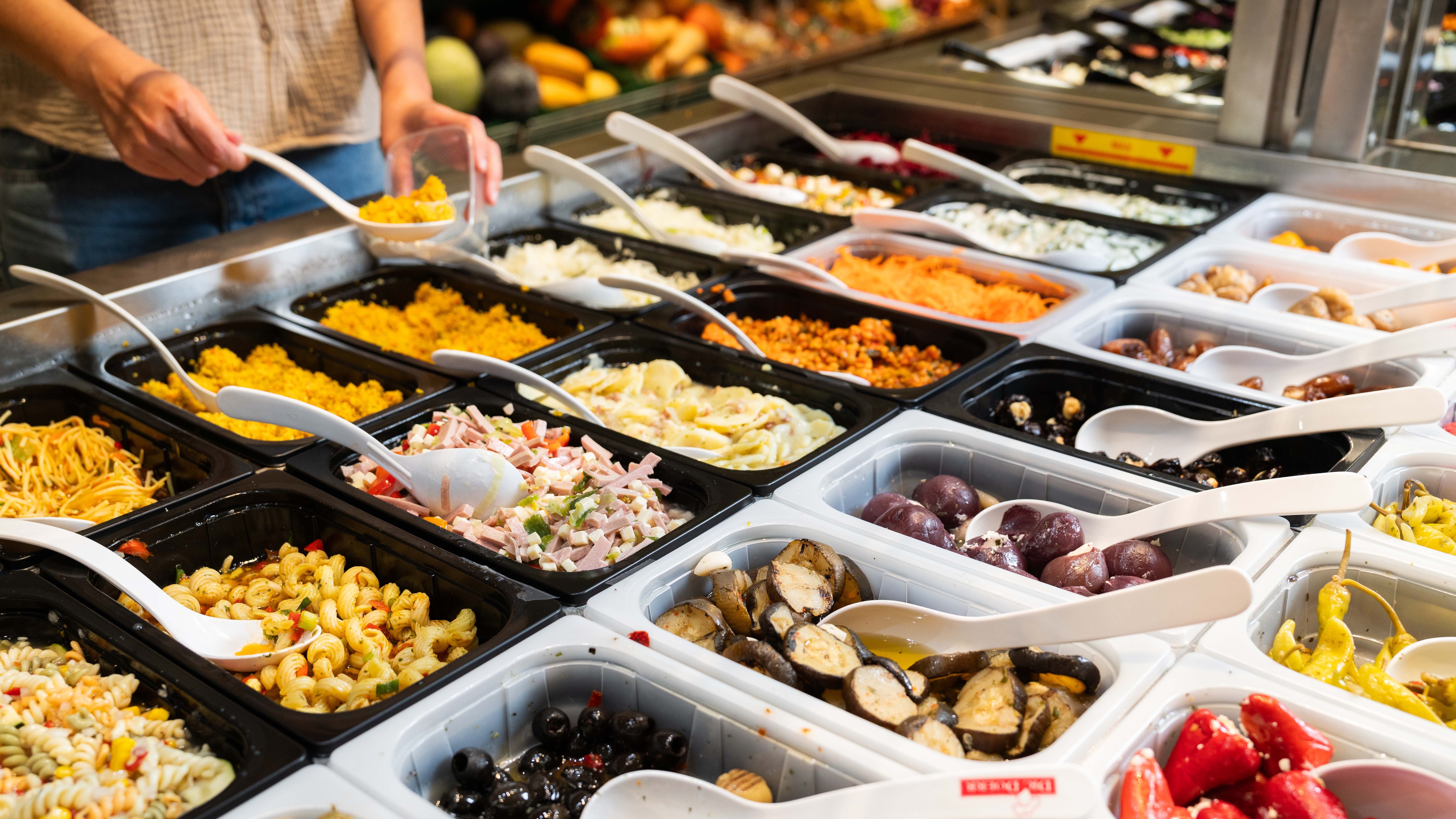 Die Salatbar mit sehr vielen verschiedenen Salatvariationen aus der sich eine Person gerade etwas in einen Becher herausschöpft.