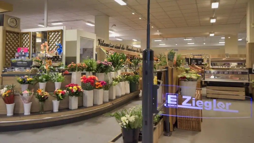 Der Eingangsbereich eines EDEKA-Marktes. Neben der Einlassschranke sind verschiedene Blumen und Pflanzen in weißen Vasen aufgestellt.