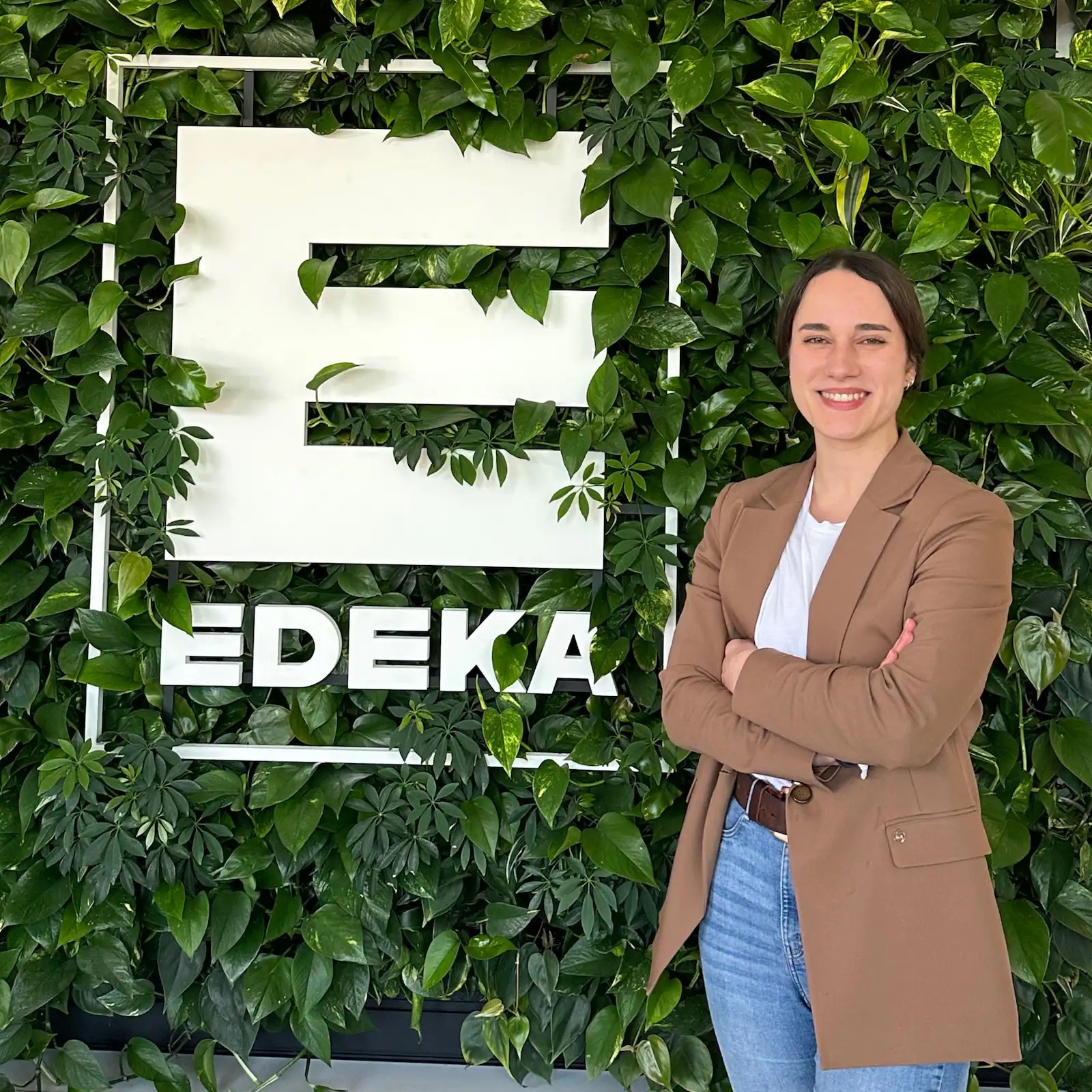 Frau in braunem Sakko und Jeans steht vor einer begrünten Fassade mit weißem EDEKA-Logo.