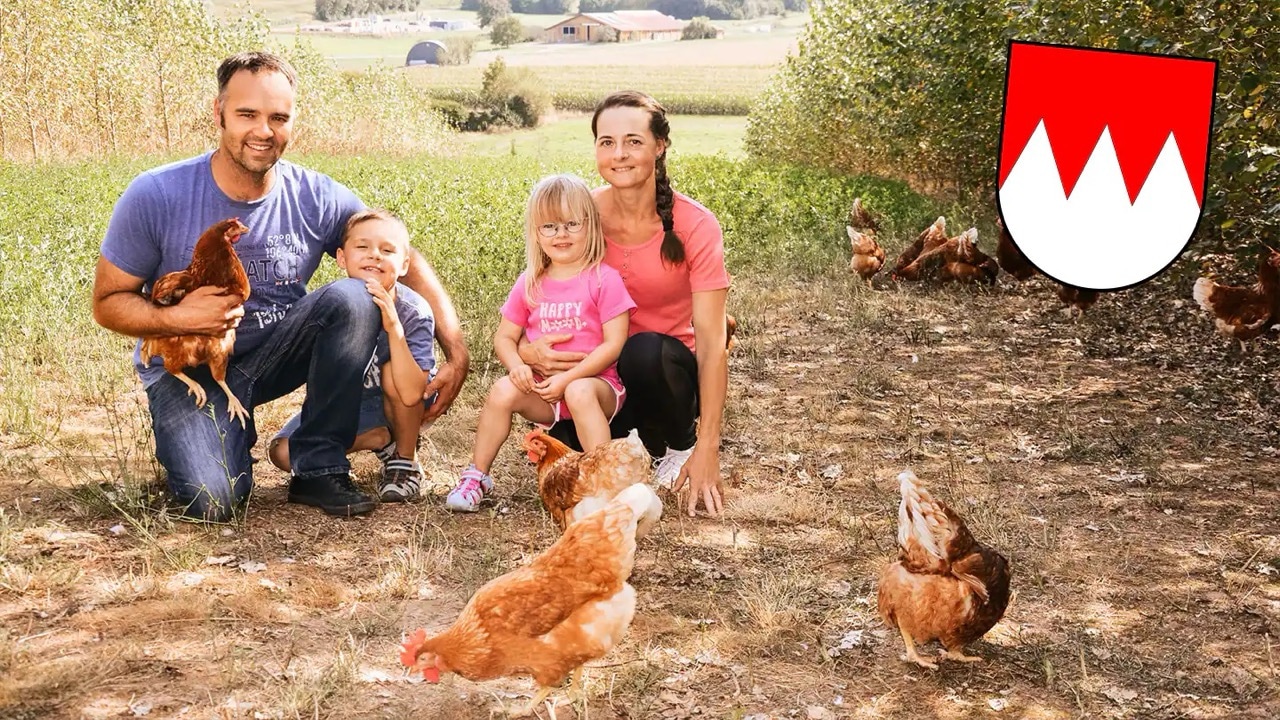 Zwei Erwachsene und zwei Kleinkinder hocken auf einer Grünfläche mit freilaufenden Hühnern. Oben rechts ein Wappen mit drei weißen Zacken auf rotem Hintergrund.