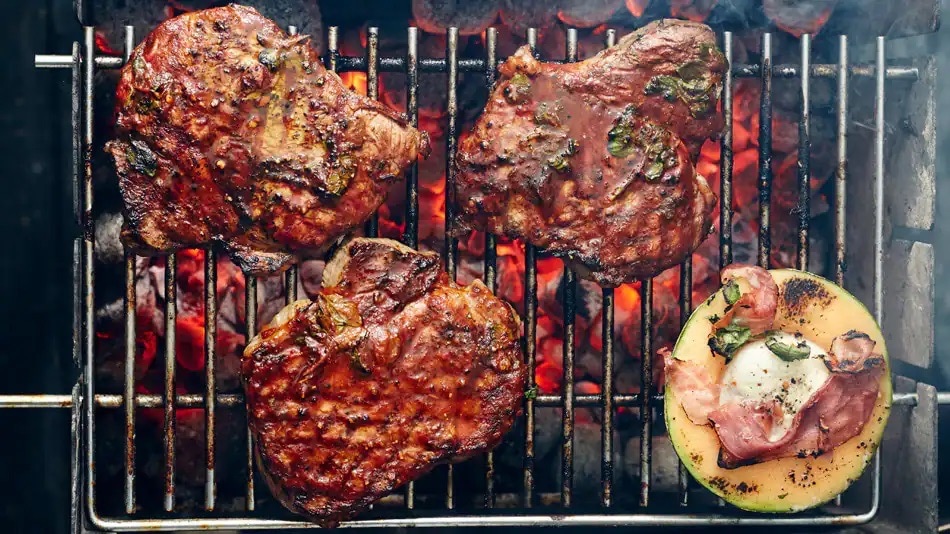 Drei Stücke Fleisch und eine Scheibe Melone auf einem Grill über glühenden Kohlen.