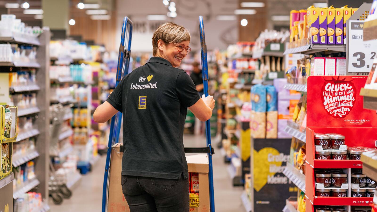 Ein Frau schiebt einen Transportroller durch den Gang eines Supermarktes. Auf dem Rücken ihres schwarzen Poloshirts steht Wir lieben Lebensmittel.